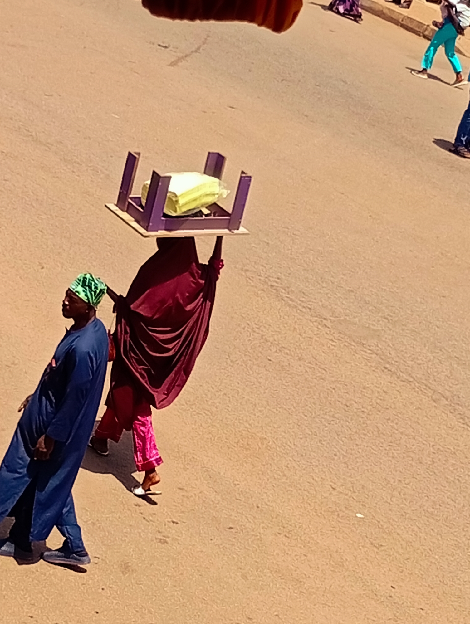 In the city of Bukuru, Nigeria, a woman balances a purple stool with a yellow package on her head. She is wearing a maroon abaya and pink trousers. To her left, a man in a blue kaftan and green headscarf walks beside her, looking forward. The ground is paved with a sandy-colored asphalt. In the background, other people are walking, including one person in teal trousers. The lighting suggests it is daytime.