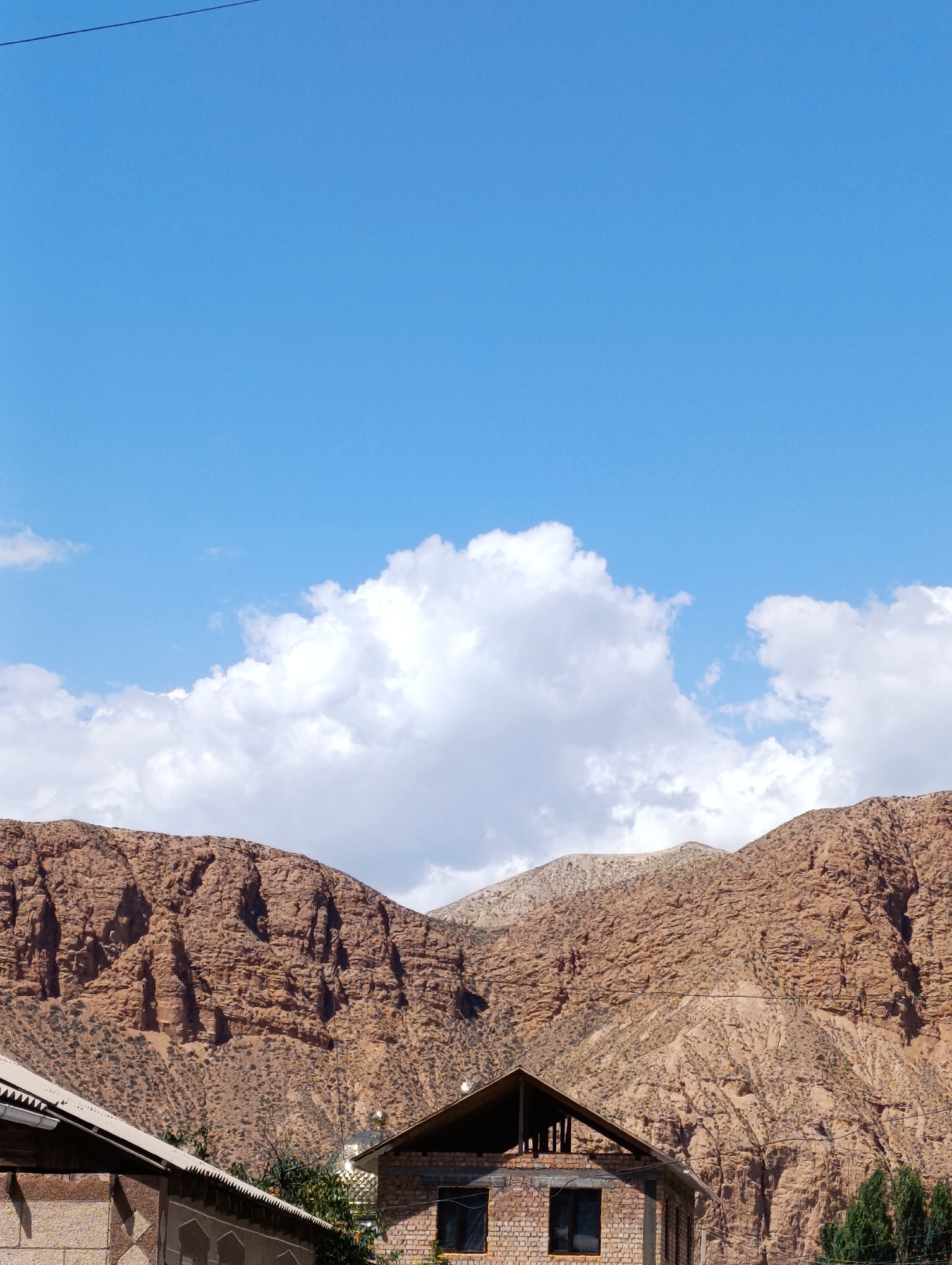 This is a landscape photograph taken in Naryn City, Kyrgyzstan, on a sunny day with a clear blue sky and scattered white clouds. The scene features a prominent, rugged mountain range in the background, characterized by its steep slopes and rocky, textured surface in shades of brown and tan. In the foreground, there are elements of human settlement. Two buildings are partially visible: one on the left with a light-colored, textured wall and a corrugated roof, and another on the right constructed of brick. This brick building appears to be unfinished, with exposed beams at the top and dark window openings. The overall impression is of a settlement nestled at the foot of imposing natural terrain.