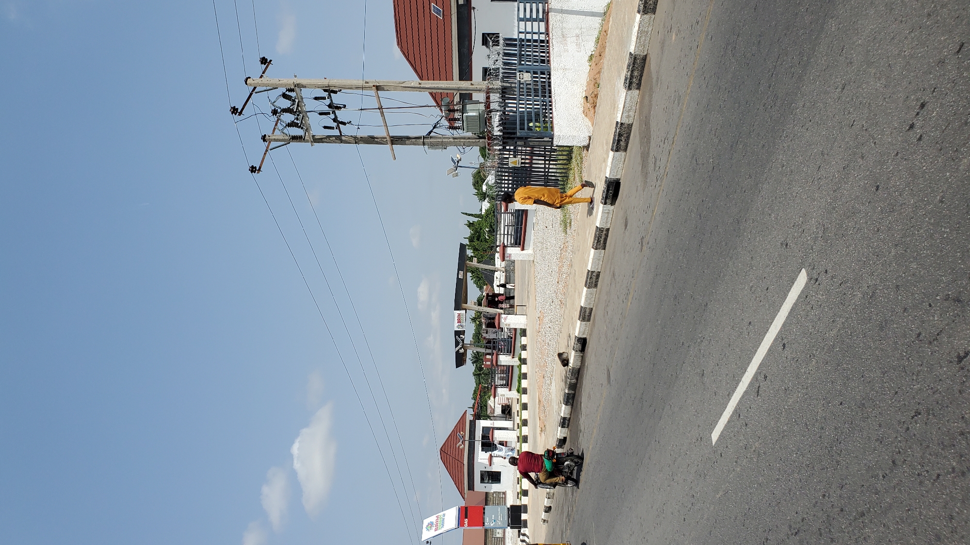 The image depicts a street scene in Jalingo, Nigeria. The focal point of the scene is a busy road with a black and white striped curb. A person dressed in an orange or yellow garment is walking along the sidewalk next to the road, with power lines and utility poles overhead. Further down the road, a gas station with several pumps is visible. A motorcycle carrying two people is in motion on the road, suggesting ongoing activity. The sky is clear and blue with a few scattered clouds, indicating a sunny day. Buildings with red-tiled roofs and modern architecture line the street, interspersed with trees and greenery. Several signs are present, including one that reads "BRIM GAS". The overall impression is one of a typical day in a Nigerian town, with everyday activities taking place along a well-trafficked road.