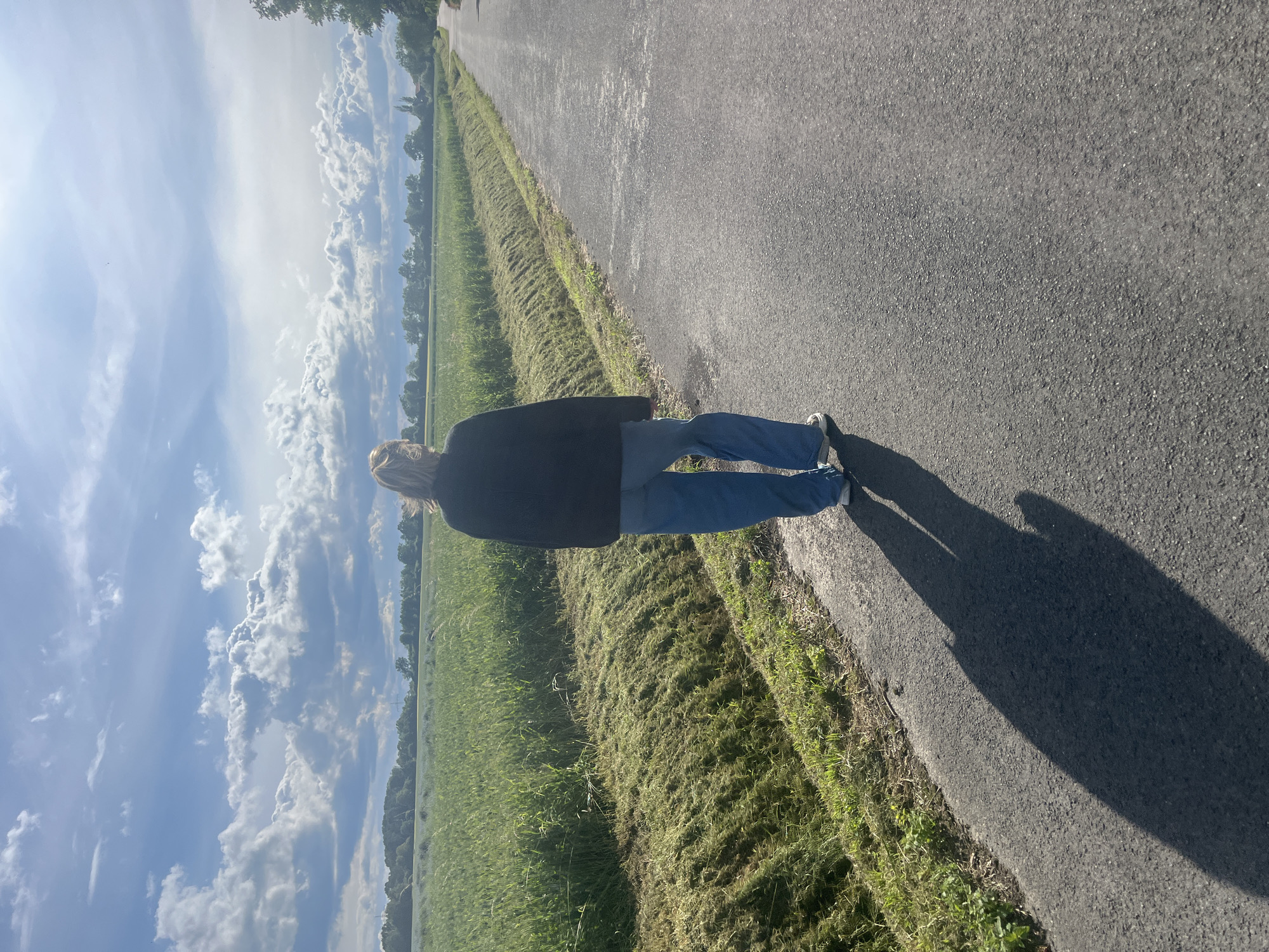 The image, oriented vertically but depicting a horizontal landscape, shows a person walking away from the camera on an asphalt road. The individual has light-colored hair, wears a dark long-sleeved top or jacket, blue jeans, and white sneakers. They are seen from the back, primarily showing their upper body and legs.
To the left of the road, a strip of recently mown grass separates the pavement from a vast green field, likely agricultural crops, which stretches towards a distant treeline under a partly cloudy sky. The sky is bright with patches of blue and numerous white, fluffy clouds.
Long, distinct shadows are cast on the road by the person, extending ahead of them (to the right in the image's current orientation). This indicates the sun is low in the sky, suggesting either early morning or late afternoon. The weather is clear and sunny with visible cloud cover. The scene evokes a rural or semi-rural setting. No discernible text is present in the image.