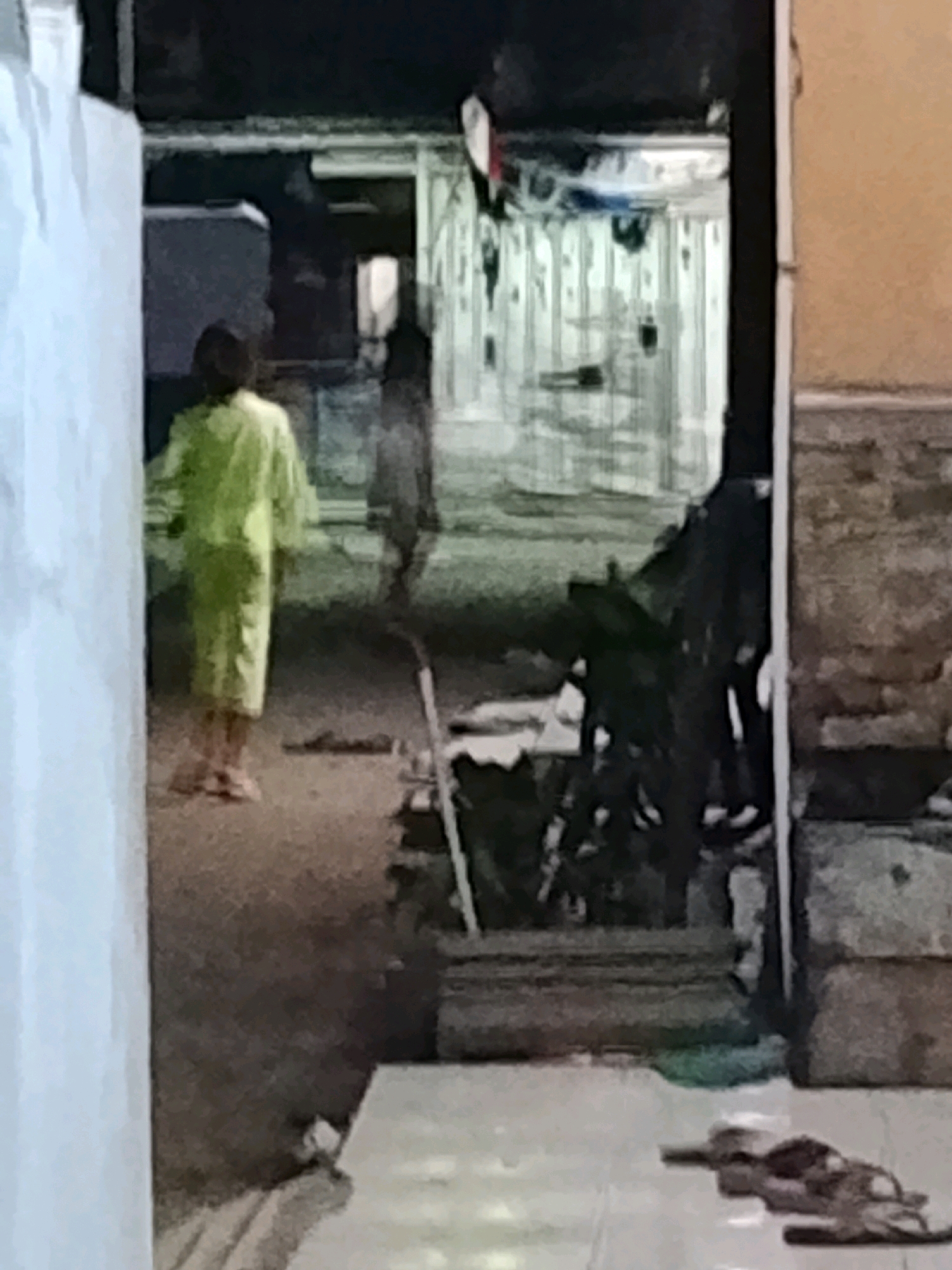 A low-resolution, dimly lit outdoor image shows a person in a bright green, long garment standing with their back to the camera. The person is in an alleyway or narrow passage between buildings. To the right of the green-garbed person, a blurred, darker figure is visible, also facing away. The ground appears to be dirt or gravel, with some debris. In the background, there are structures that appear to be part of a dwelling or workshop, with what looks like laundry or fabric hanging. The overall scene suggests a residential or utilitarian area in a city like Ujungrusi, Indonesia, possibly at dusk or night due to the low light and indistinct details.
