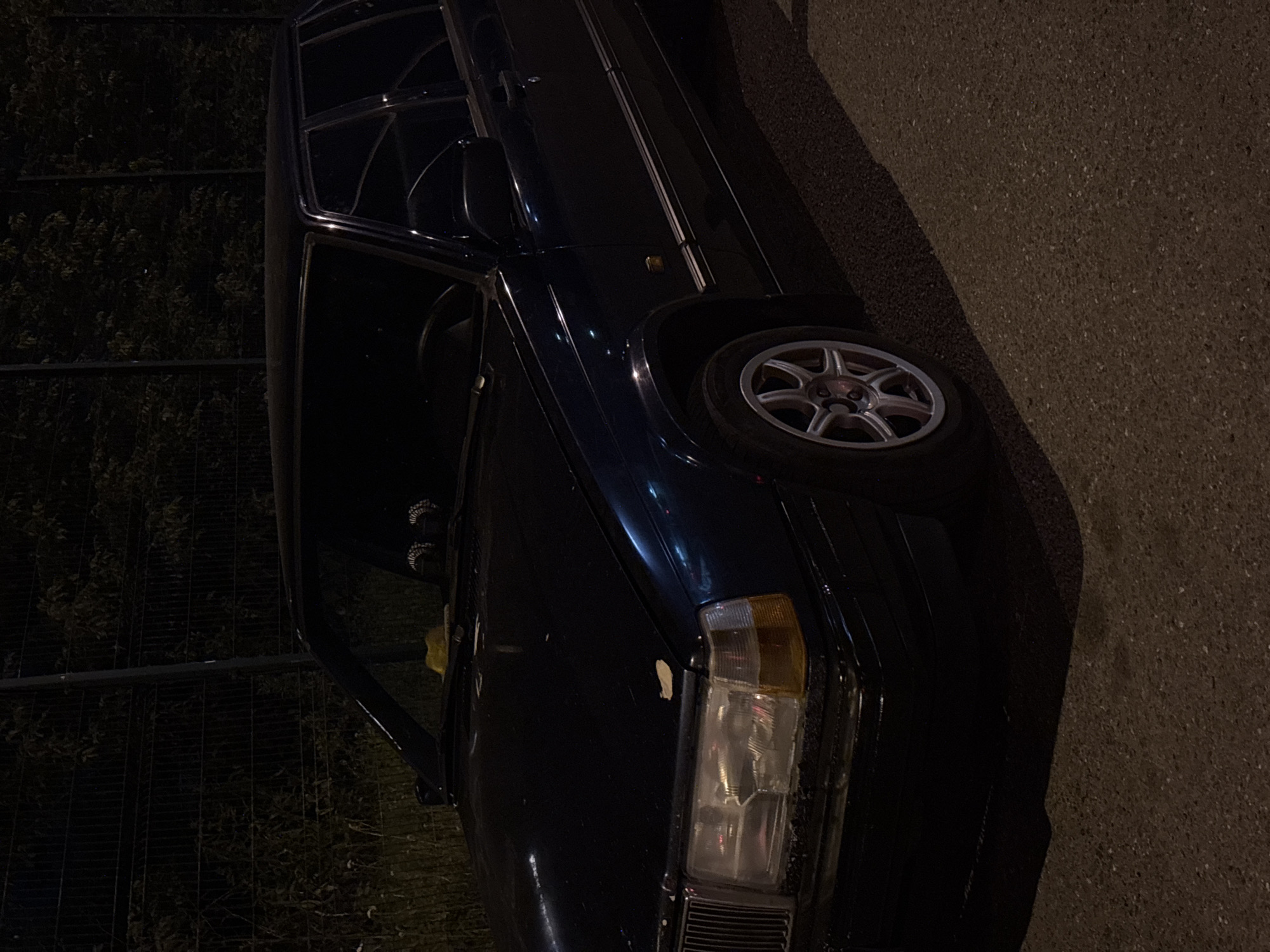 An aged, dark blue Volvo sedan is parked on asphalt, possibly in Novokuznetsk, Russia, at night. The car's headlights are visible, and the scene is dimly lit by an unseen light source, casting shadows. A chain-link fence and dark foliage form the backdrop. The car appears to be in decent condition, with some wear and tear visible, particularly around the headlights and bumper. The pavement is textured and appears to be a public road or parking area. No people are visible in the image.