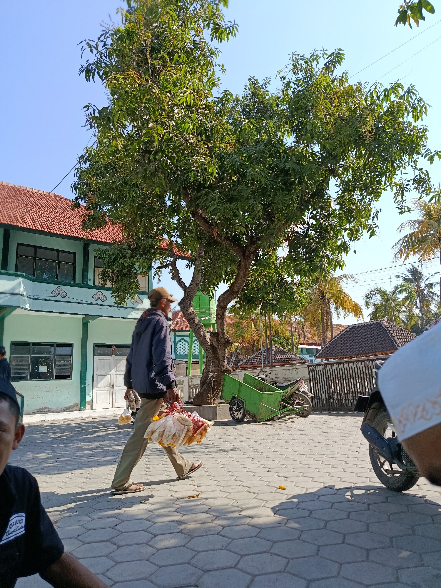 In Ramban Wetan, Indonesia, a man walks across a paved courtyard in front of a light green building with a red tiled roof. He is wearing a dark jacket, light brown pants, a baseball cap, and sandals, and carries several bags filled with food items. A large tree with green leaves stands to his right, and a green cart and a motorcycle are visible behind him. In the foreground, a person wearing a black t-shirt and another person wearing a white hat are partially visible. The sky is clear and blue.