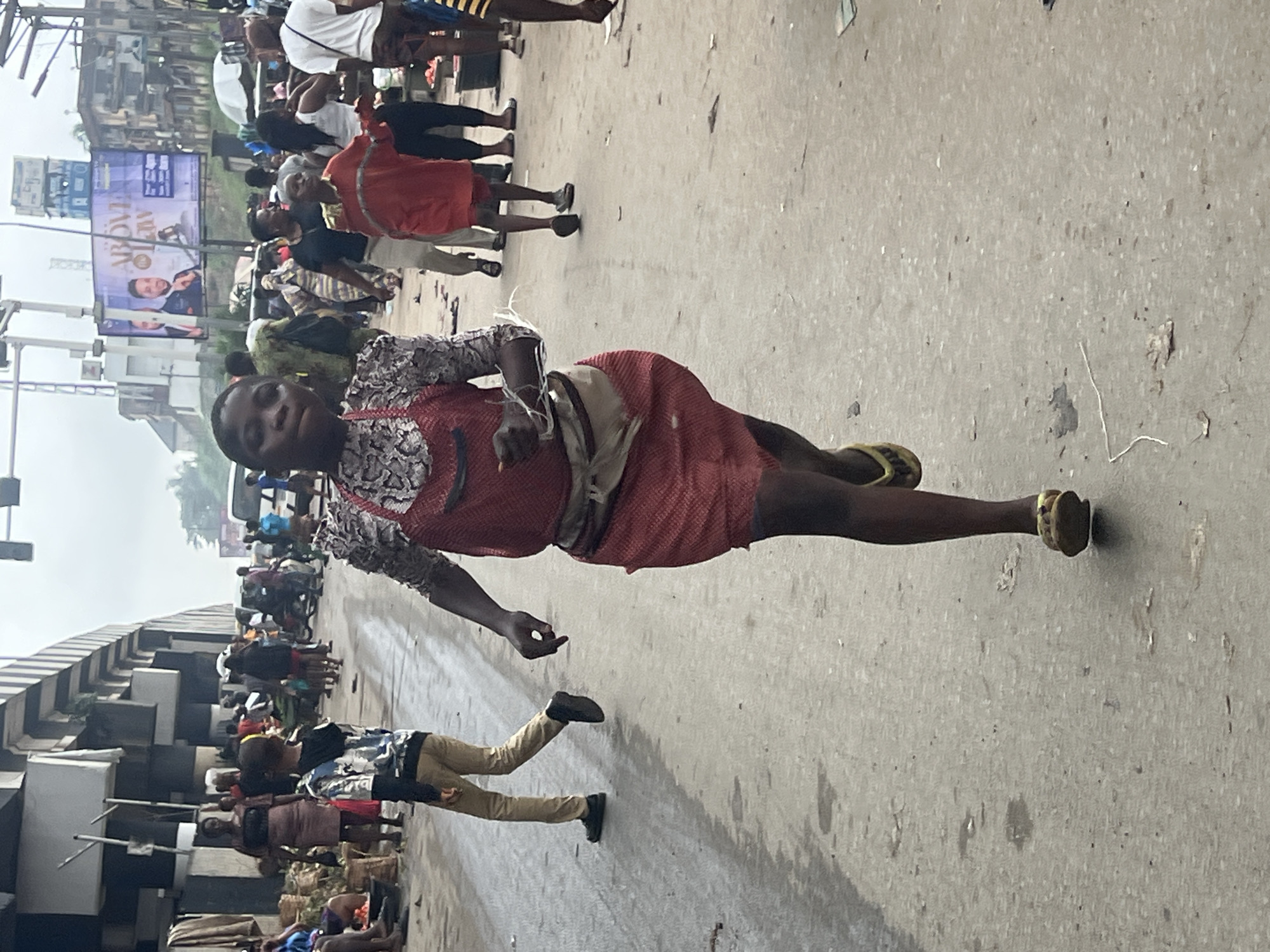 In the city of Nkpor, Nigeria, a young girl in a red patterned dress and white top is running towards the camera on a paved road. She has a light-colored sash around her waist, and is wearing yellow sandals. The road is wet and has some debris scattered on it. In the background, a crowd of people and market stalls can be seen. A large billboard advertising "ABOVE ALL LAW" is also visible. A young boy in a grey and white patterned shirt and khaki pants is walking away from the camera on the left side of the road. Other individuals in various clothing are scattered throughout the scene. The atmosphere appears to be that of a busy market or street.