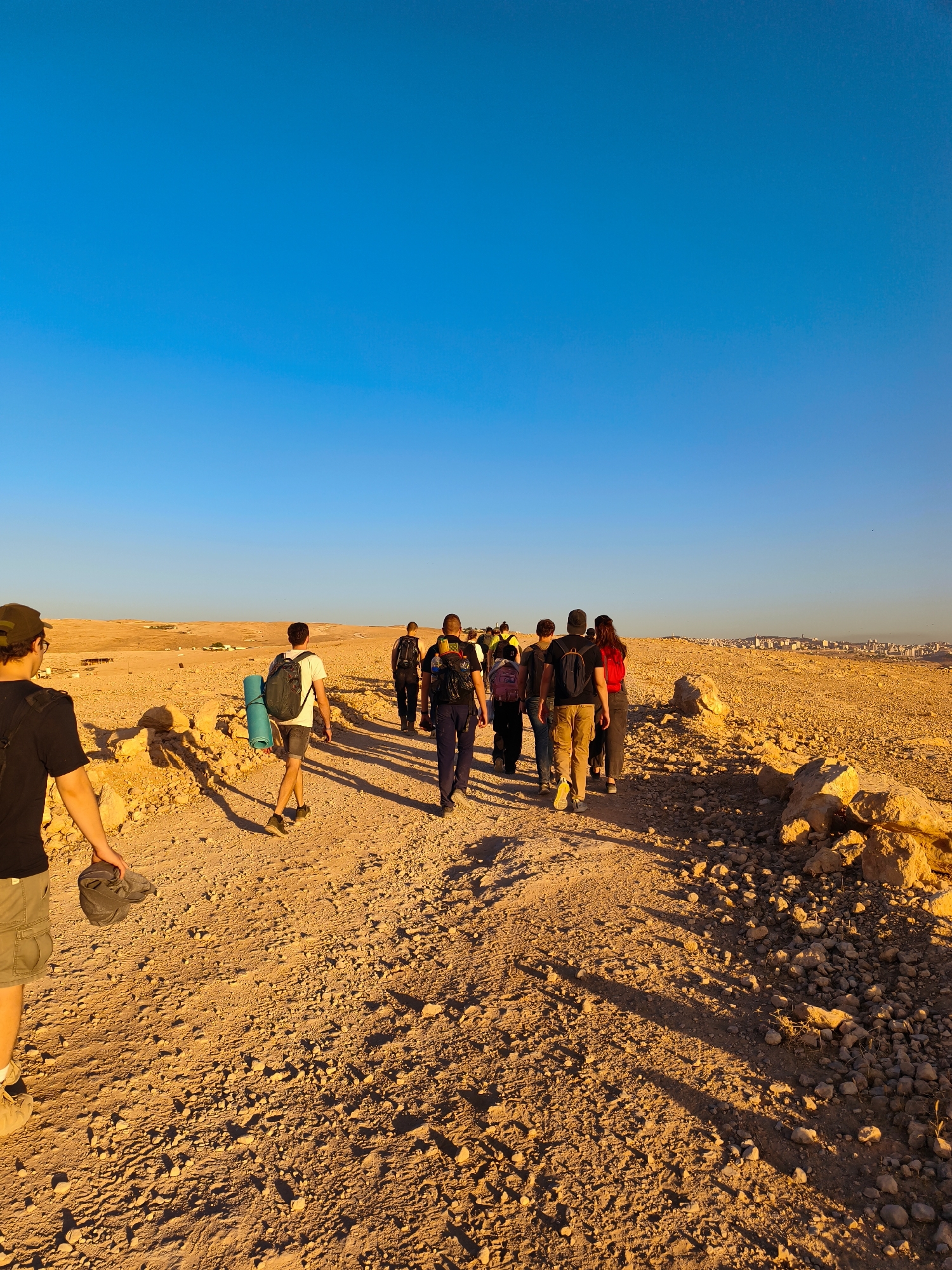 A group of approximately ten individuals are walking along a dirt path in a rocky, arid landscape under a clear blue sky. The sun is low, casting long shadows from the people and rocks. The individuals are wearing casual clothing suitable for hiking and several have backpacks, with one person carrying a rolled-up sleeping pad. In the distance, a city skyline is visible on a horizon line dotted with buildings. The terrain is characterized by dry earth, scattered rocks, and sparse vegetation. The scene suggests a hike or expedition in a desert or semi-arid region.