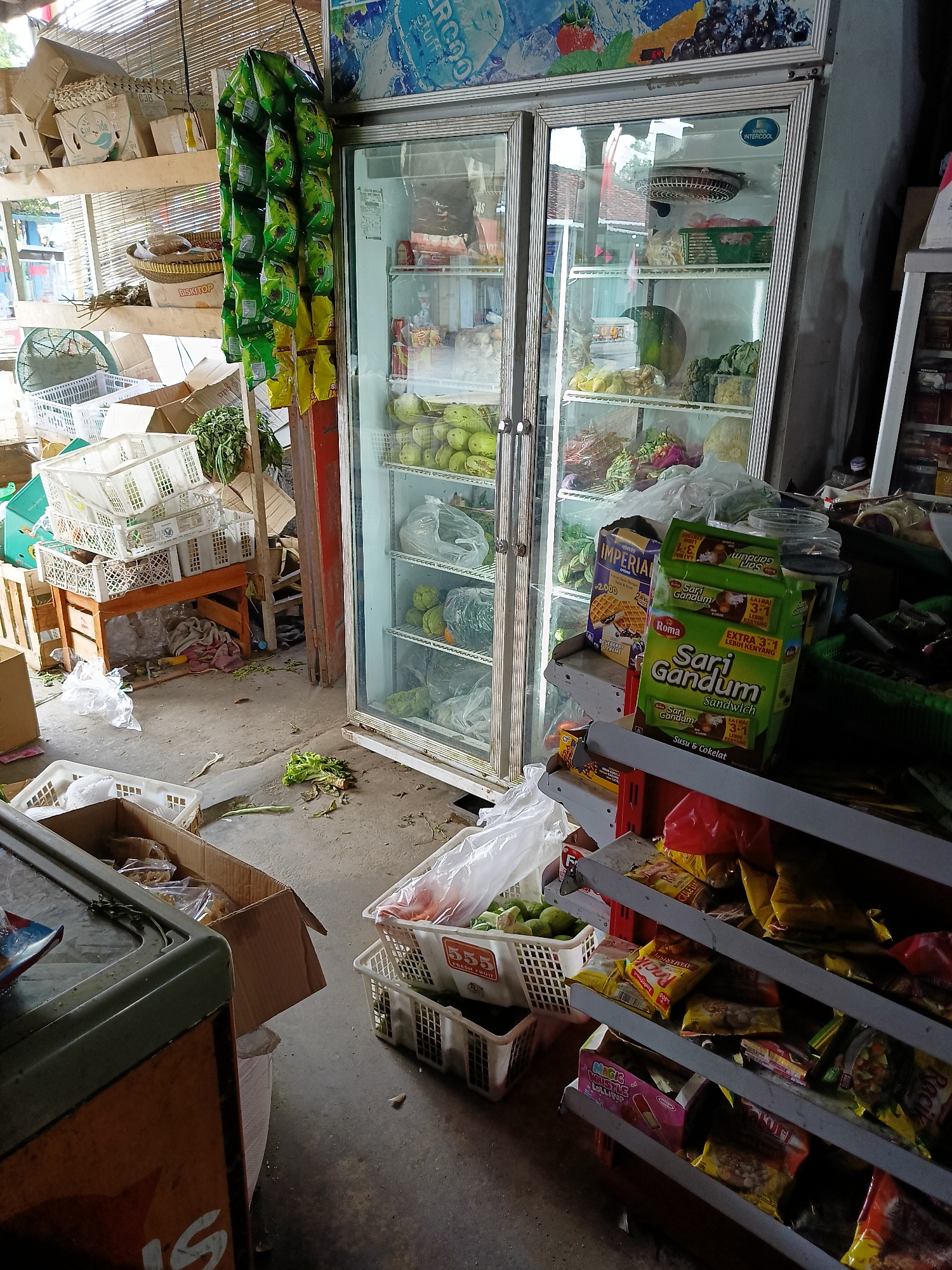 This is a medium-shot, eye-level photograph of the interior of a small, cluttered convenience store in Sukorini, Indonesia. The store is filled with various items, including a large glass-door refrigerator stocked with produce like guavas, broccoli, and leafy greens. Baskets of fruits and vegetables are also visible on the floor and shelves, some containing packaged goods. Several cardboard boxes are stacked around the store, and some shelves display packaged snacks and food items. A collection of green and yellow snack packets hangs from the ceiling near the refrigerator. The floor is concrete and appears somewhat dirty, with scattered debris. There are no people visible in the image. The overall impression is one of a busy, somewhat disorganized, local shop.