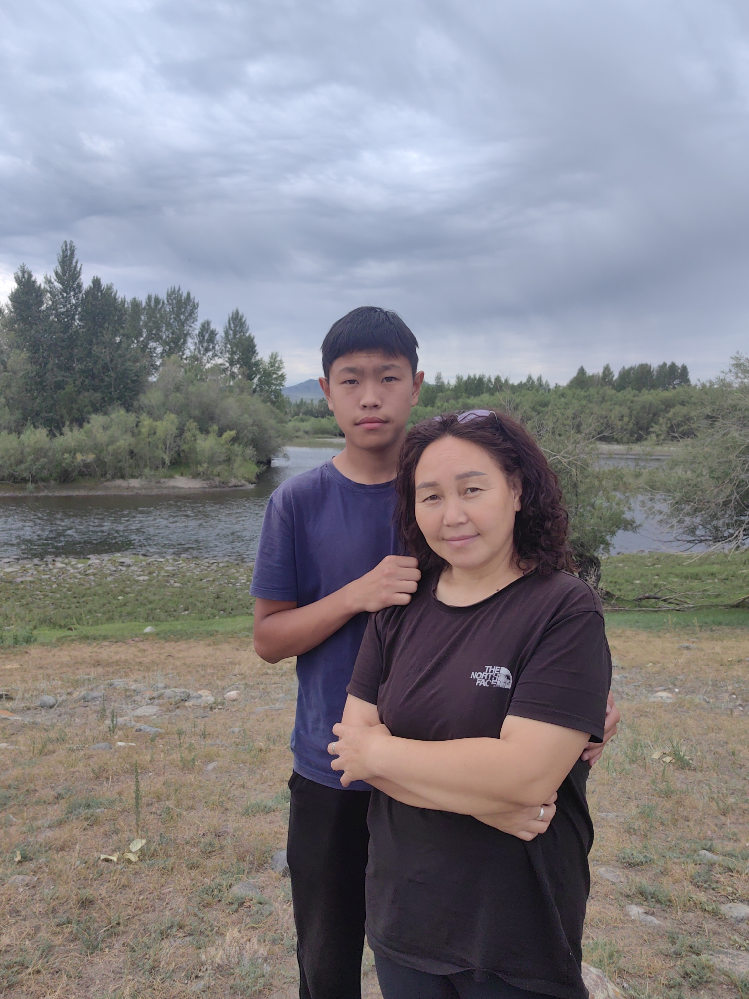 A medium shot shows a woman and a teenage boy standing outdoors. The woman is in the foreground, slightly to the right of center, and is looking at the camera with a slight smile. She has curly dark hair and is wearing a black t-shirt with a white logo. The teenage boy is standing behind and to the left of the woman, with his right arm around her shoulders. He is looking directly at the camera with a neutral expression and is wearing a dark blue t-shirt. They are positioned in front of a body of water, likely a river, with a tree-lined bank on the opposite side. The sky is overcast with gray clouds. The ground in the foreground is covered with dry grass and scattered rocks. The scene appears to be in a natural outdoor environment.