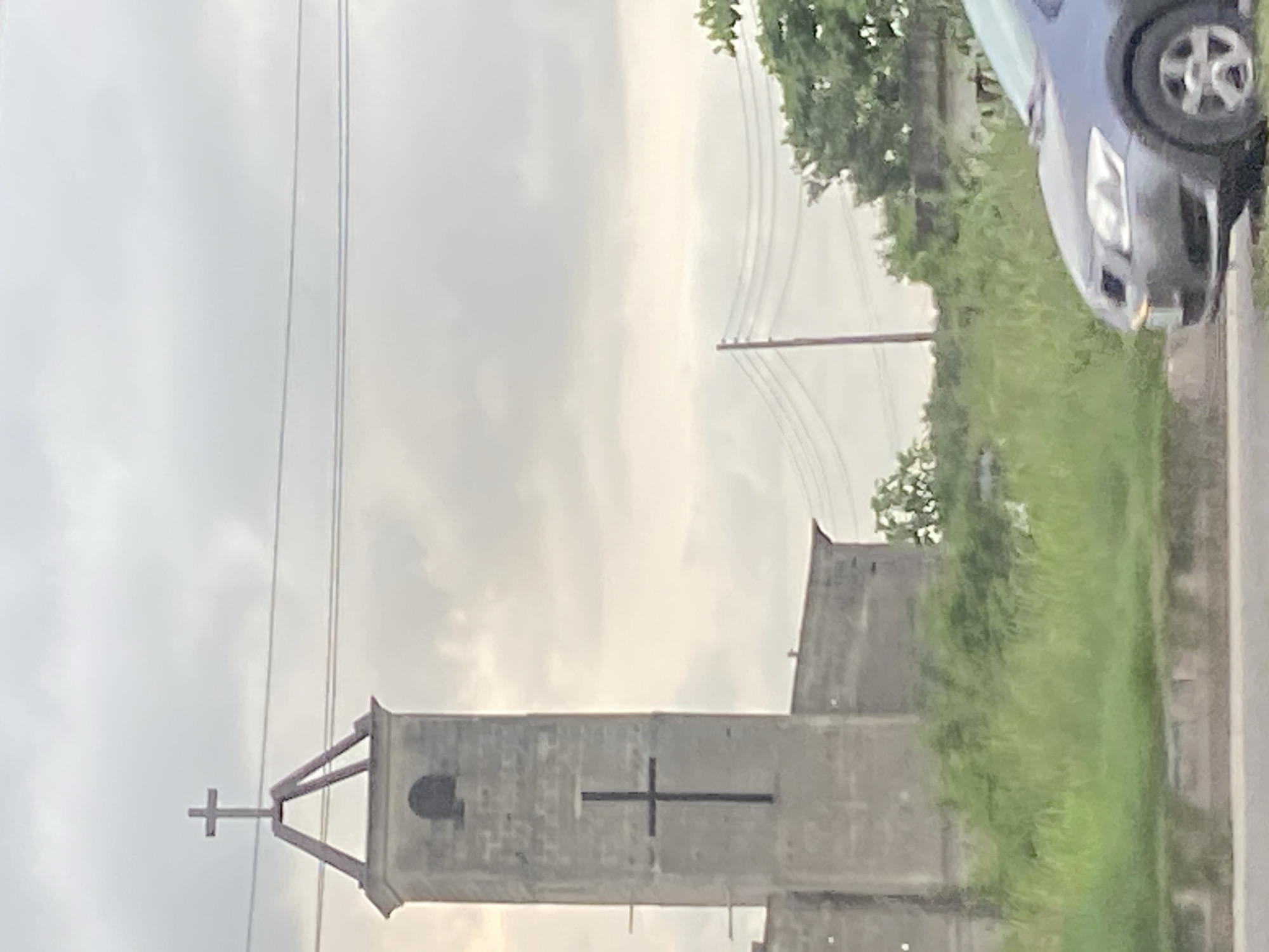 The image depicts a view of a church structure in Ikorodu, Nigeria, with a prominent cross on its steeple and another large cross on its facade. The church appears to be constructed of concrete blocks. Adjacent to the church, there is a substantial amount of overgrown green vegetation. In the foreground, a grey car is partially visible, parked on what appears to be a roadside. Overhead, power lines stretch across the frame, suggesting an urban or semi-urban setting. The sky is overcast with clouds, indicating a diffused light condition.
