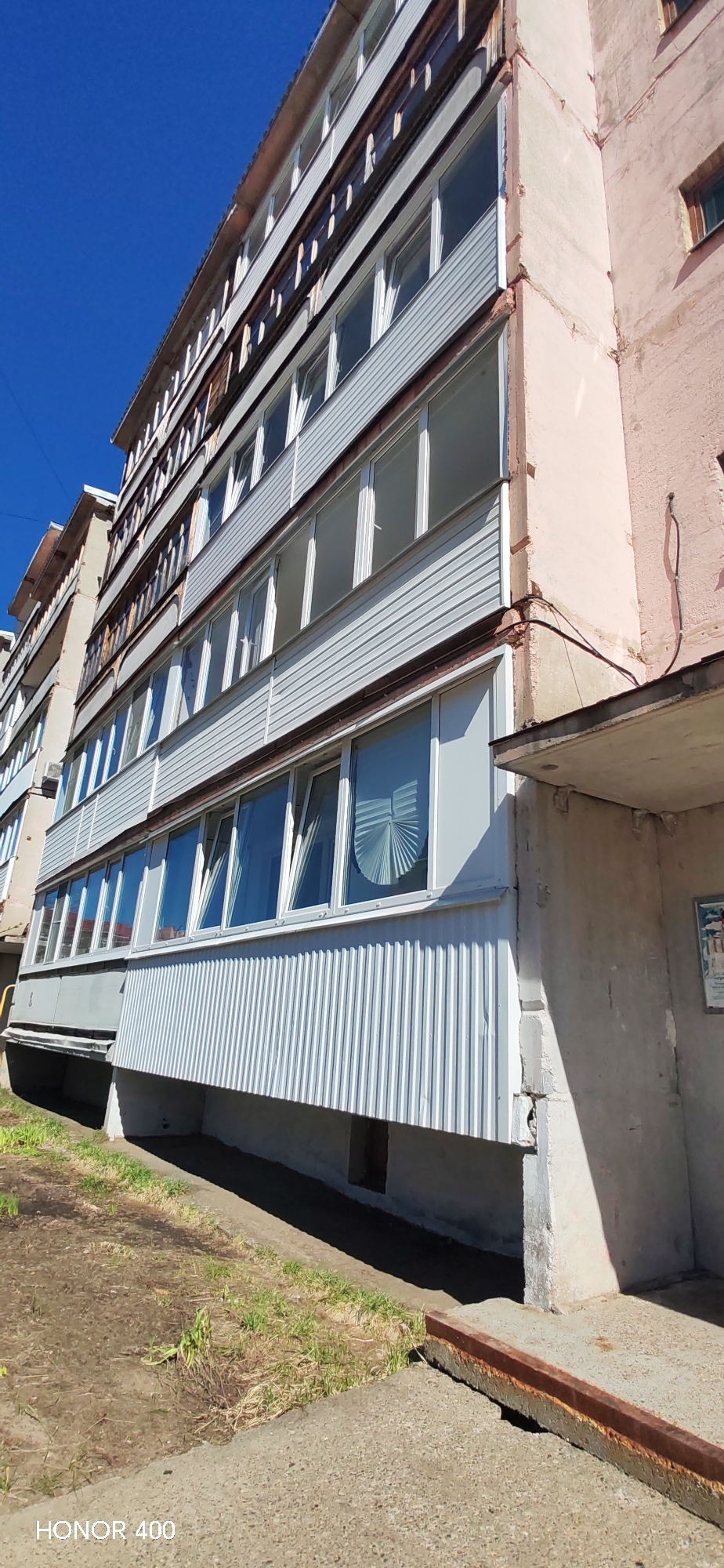 The image depicts an apartment building in Noviy, Russia. The building has multiple floors with balconies, some of which are enclosed with white corrugated metal. The building's facade appears to be in a state of disrepair, with visible wear and tear. The sky is clear and blue. The foreground shows a patch of bare ground with some grass. The image has "HONOR 400" imprinted on the bottom left.