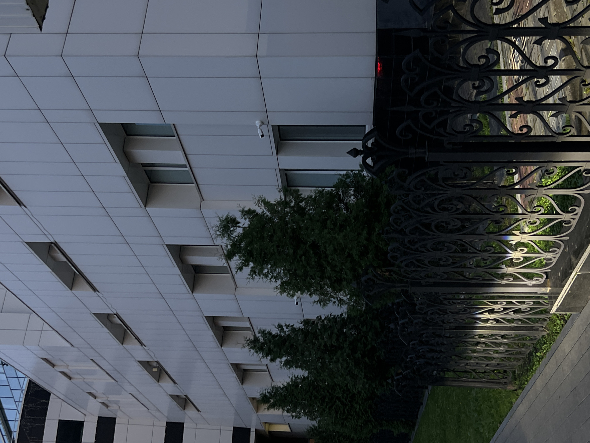 The image shows the exterior of a modern, multi-story building with a white, tiled facade. Several windows are visible, some recessed with visible interior structures. A security camera is mounted on the building's exterior. In front of the building, there is a neatly trimmed green lawn and a row of tall, dark green evergreen trees. A decorative black metal fence runs along the front of the property, with ornate scrollwork. The ground in front of the fence appears to be a paved walkway. The lighting suggests it is either dawn or dusk, with artificial lights casting distinct patterns on the fence and walkway. The building's architecture suggests it is an institutional or commercial building. While the image does not explicitly state the location, the style of architecture and fencing are consistent with urban environments in Russia.