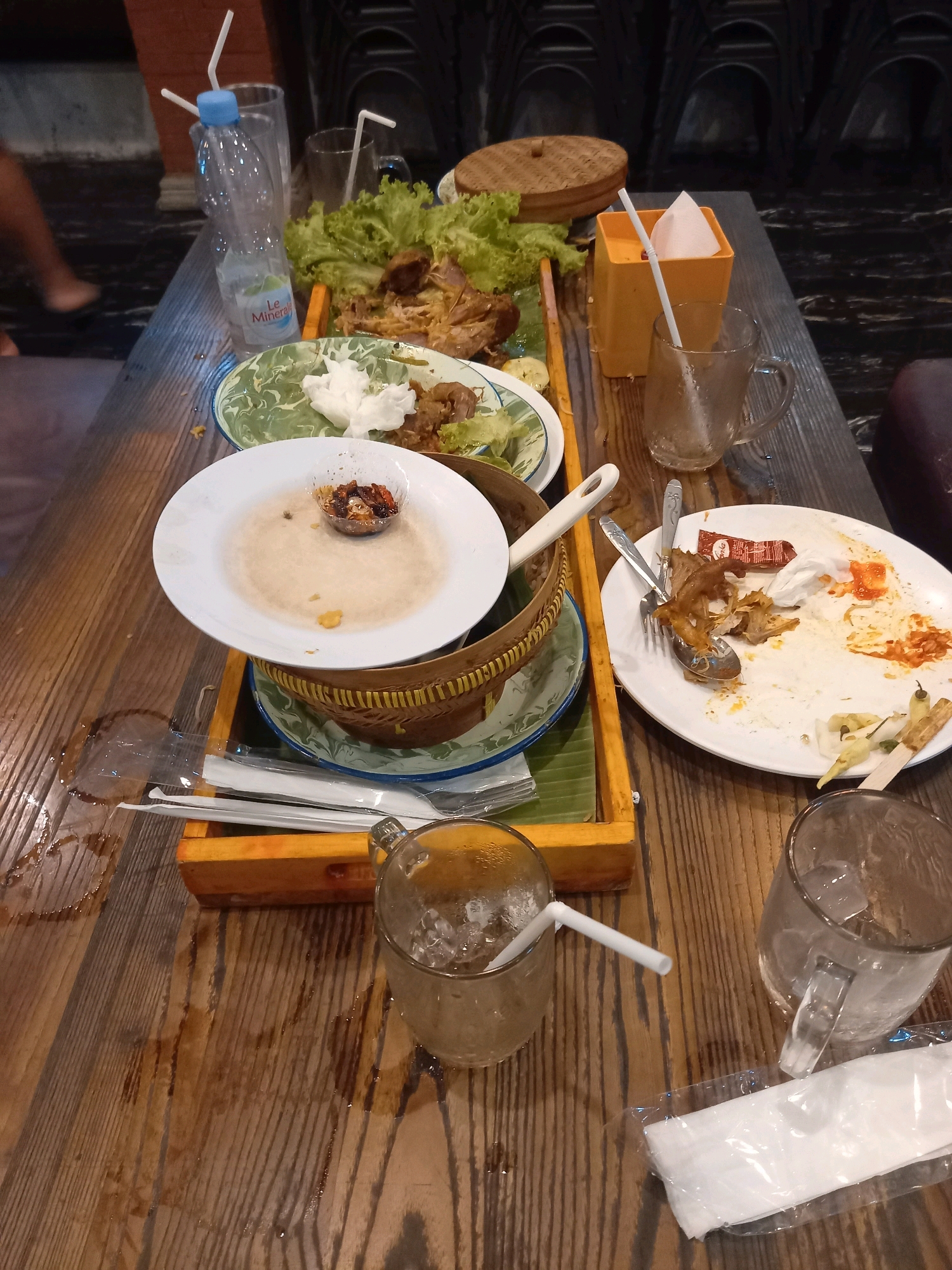 The image depicts a table at what appears to be a restaurant in Kepanjen, Indonesia, shortly after a meal. The table is made of dark wood and has a wet, slightly sticky appearance, suggesting spills. The setting is dimly lit, with a background of dark, shadowed seating and some decorative arches.
On the table, remnants of a meal are evident. Several used plates are present, some with leftover food, bones, and sauces. A white plate in the foreground has a small bowl of dark red chili sauce and a few crumbs. Another white plate on the right is heavily soiled with remnants of meat, rice, and red sauce, along with a crumpled napkin and a few small green chilies on skewers. There are also several empty drinking glasses, some containing ice cubes and straws, and one glass has a handle.
A plastic bottle of "Le Minerale" drinking water is also on the table, near a glass. Other objects include a woven bamboo basket, a wooden tray holding cutlery wrapped in plastic, and a yellow dispenser for napkins, which contains a folded white napkin and a straw. A person's bare leg and foot are partially visible in the upper left corner of the image, indicating their presence at the table. The overall impression is one of a meal that has recently concluded, with the table left in a state of moderate disarray, typical of a busy dining establishment.