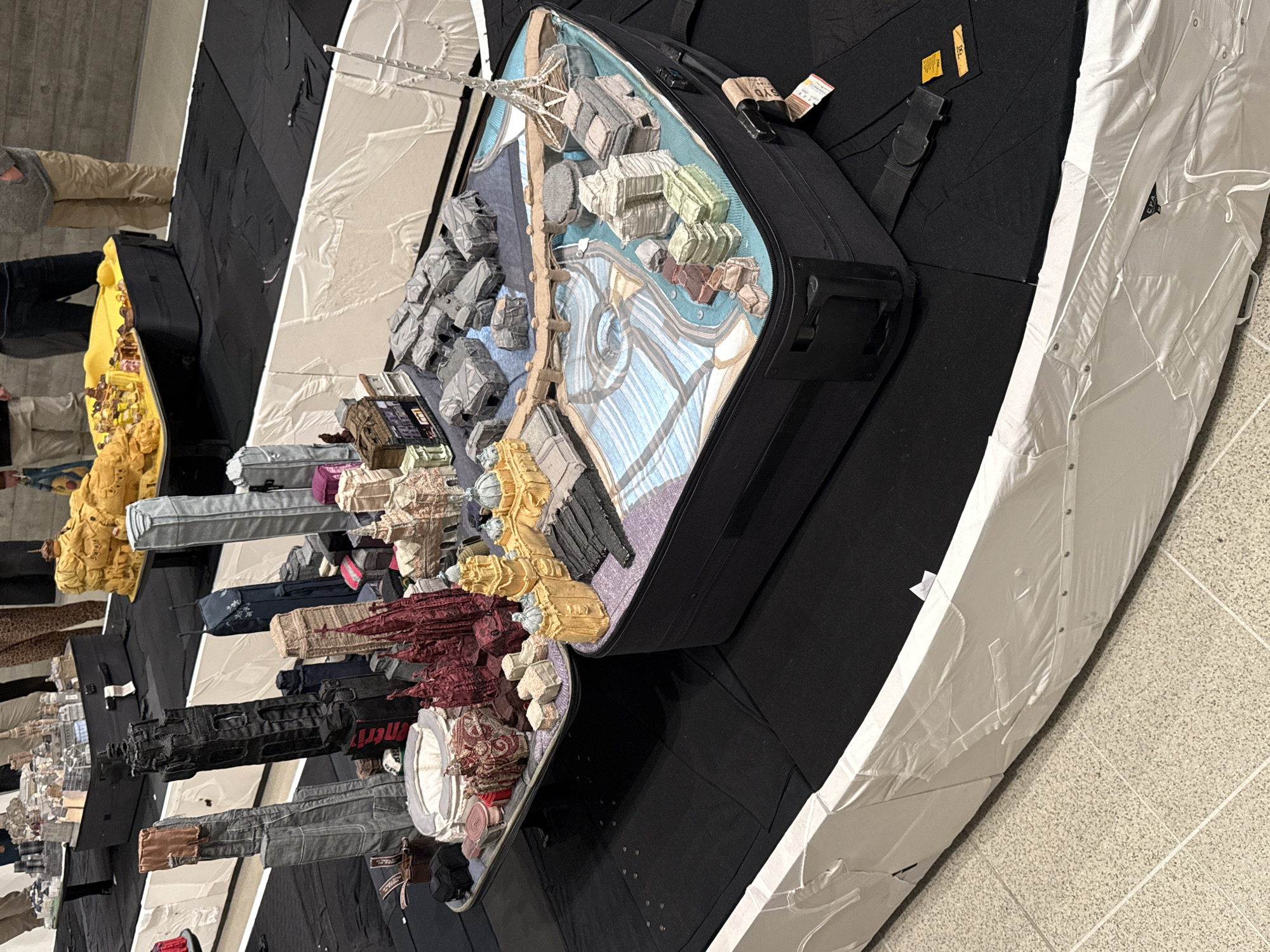 The image shows an art installation constructed within a suitcase, depicting a city skyline or a map of an urban area. The buildings and structures appear to be crafted from fabric and various craft materials, suggesting a tactile and handmade approach. The art installation appears to depict a miniature version of the city of London, given the inclusion of the London Eye or a similar Ferris wheel. The suitcase rests on a platform covered in black and white material, giving the impression of a stage or display area within an exhibition space. The environment seems to be indoors, likely within an art gallery or museum setting. Other suitcases with fabric buildings are visible on the left of the suitcase.