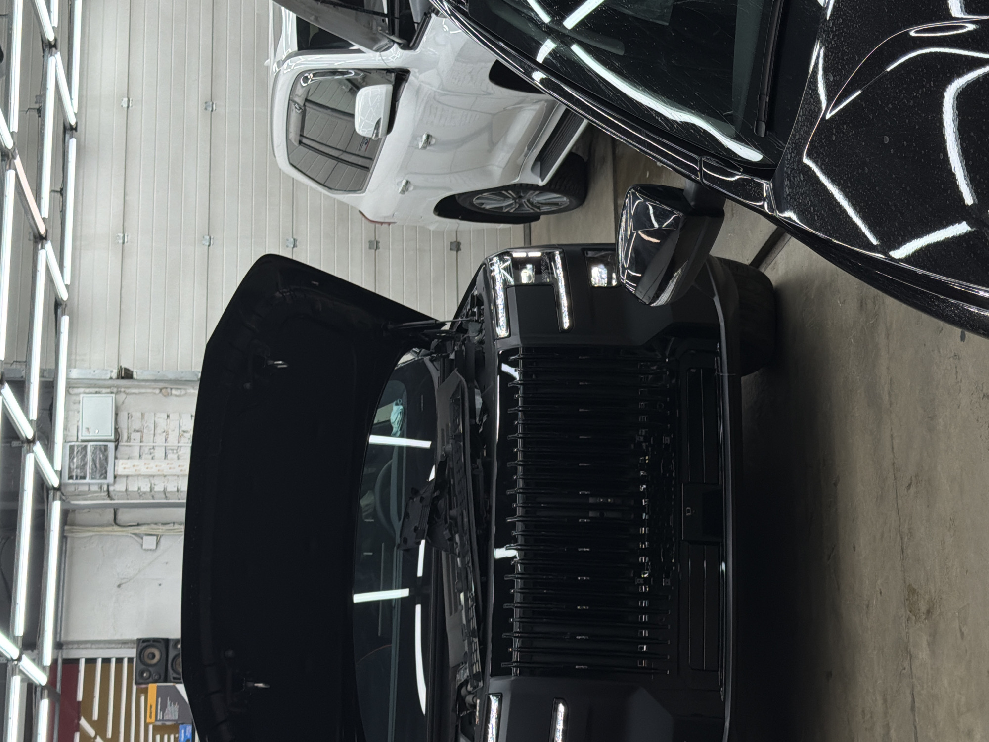 The image shows two cars parked in an indoor space that appears to be a car detailing or repair shop. One car is white and the other is black. The hood of the black car is open. The location could be Krasnoyarsk, Russia, but there is no evidence to confirm the location. There are no people visible in the image. Fluorescent lights are visible on the left side of the image.