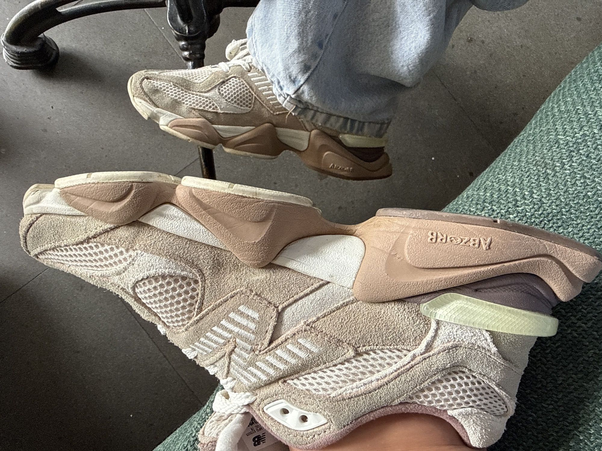 A close-up shot focuses on two New Balance running shoes. One shoe is tan, featuring white stripes and a prominent "ABZORB" imprint on its brown sole. The other shoe, partially visible under a leg clad in light-wash denim jeans, is also tan, with a brown sole. Both shoes have a mesh pattern and appear to be well-worn. The background includes a dark grey floor, a portion of a black metal chair, and a textured green surface, likely part of a chair or cushion. The location of Ubud District, Indonesia, is not evidenced by the image.