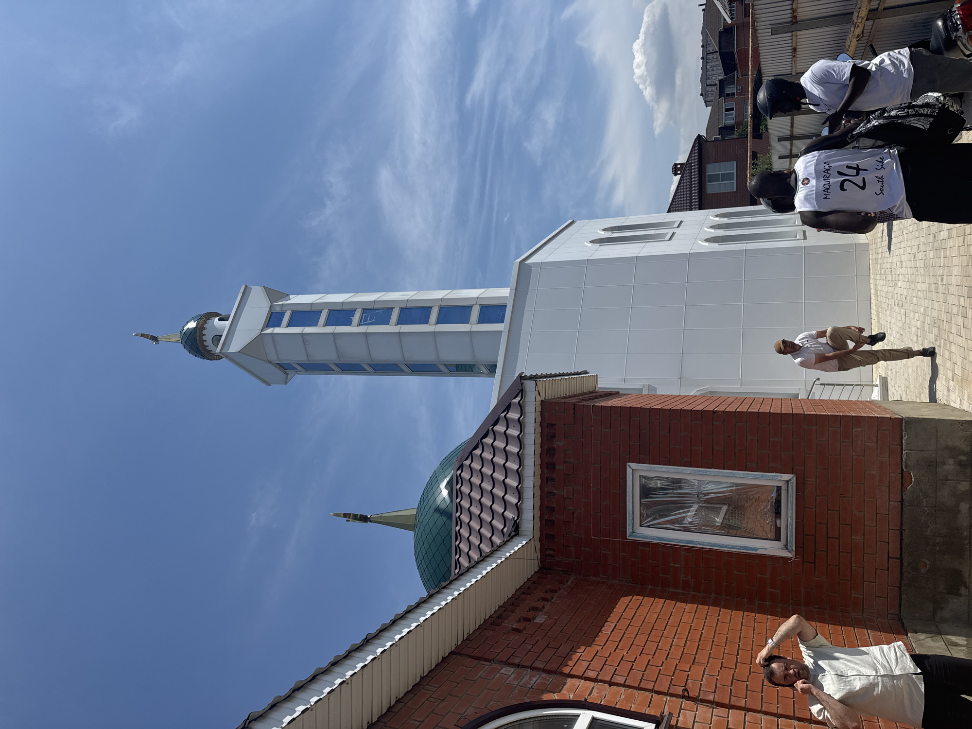 The image shows a mosque in Novaya Adygeya, Russia, under a clear blue sky with scattered clouds. The mosque features a green dome topped with a gold crescent, and a tall, white minaret with blue-tinted windows. A red brick building, possibly part of the mosque complex, is to the left, with a brown tiled roof.
In the foreground, three men are visible. One man, wearing a light-colored shirt and dark pants, is standing near the brick building, holding a phone to his ear and looking towards the camera. Another man, wearing a white tank top with the number "24" and "MAQUIRAGA SOUTH SIDE" printed on it, is facing away from the camera, talking to a third man in a white t-shirt and helmet. A motorcycle is parked to the right. Another man, wearing a light polo shirt and khaki pants, is standing with one leg raised, looking towards the mosque. The ground is paved with interlocking bricks. In the background, residential buildings and fences are visible.