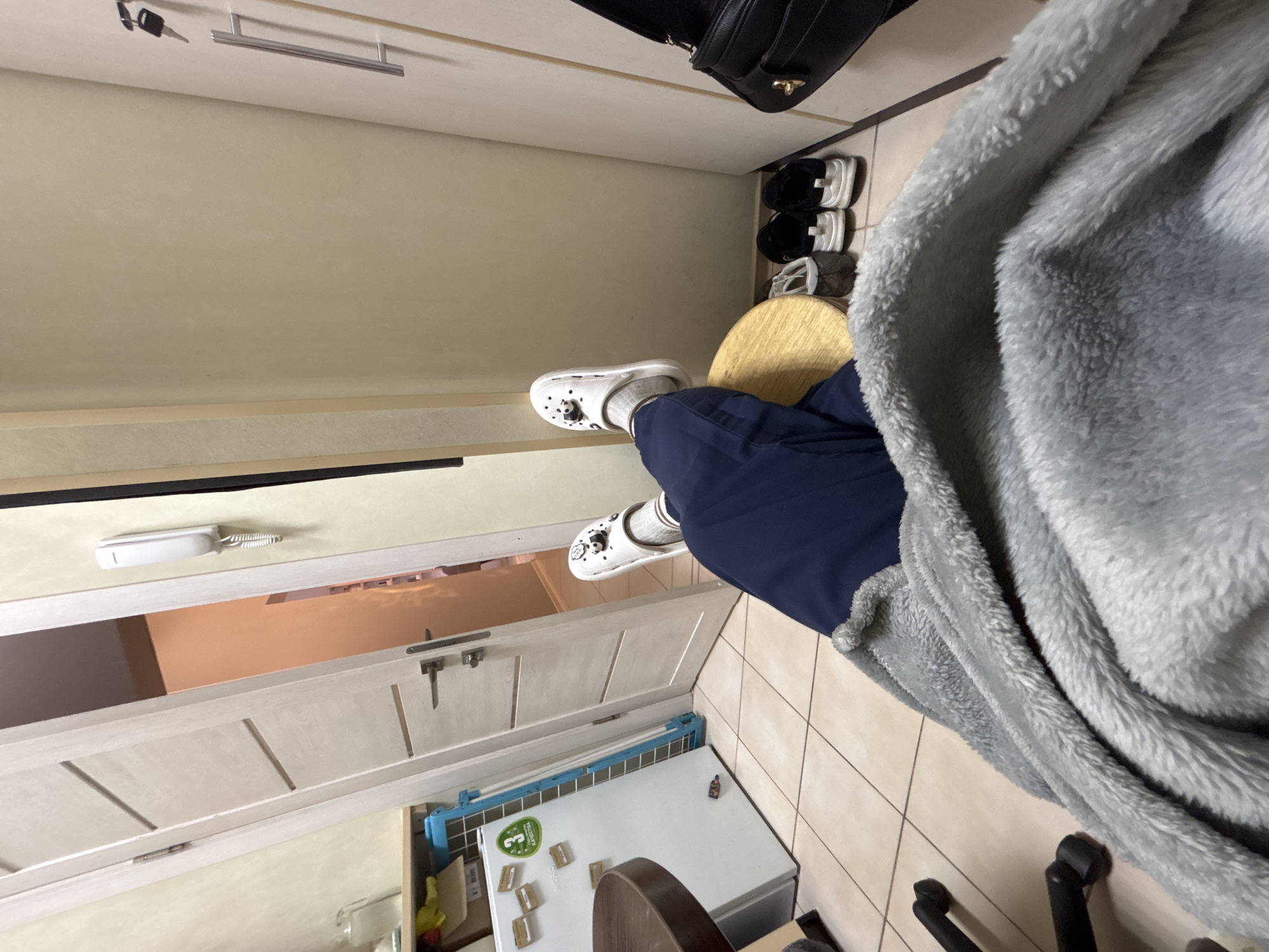 A first-person perspective photo reveals a tiled floor room in Unknown, Unknown. The individual is seated on a round wooden stool, legs covered in blue fabric, feet adorned with white Crocs and gray socks. A fuzzy gray blanket partially obscures the view. Adjacent is a white refrigerator, affixed with a collection of magnets and stickers. A white door, partially open, leads to an adjacent room. On the wall, an intercom system is installed. Further in the background, shoes are arranged, and a black purse rests. A set of keys is visible on a higher surface.