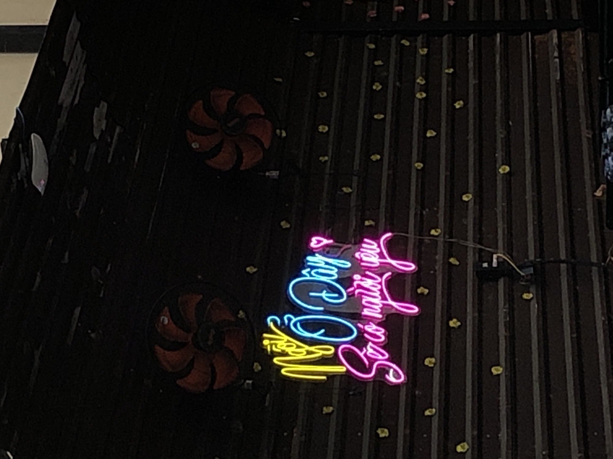 This is a medium shot, eye-level, exterior photograph of a closed storefront with a corrugated metal roll-up door. Two red, circular ventilation fans are mounted above the door. A neon sign is displayed on the door, with the text "Ngồi Ở Đây So có người yêu" written in yellow, blue, and pink. The sign is illuminated, casting light on the dark metal door, which is decorated with small yellow, flower-shaped stickers. A black electrical wire hangs down from the sign, connected to a black electrical box mounted on the door. The scene appears to be in Đà Nẵng, Vietnam, likely a street-level establishment. No people are visible in the frame.