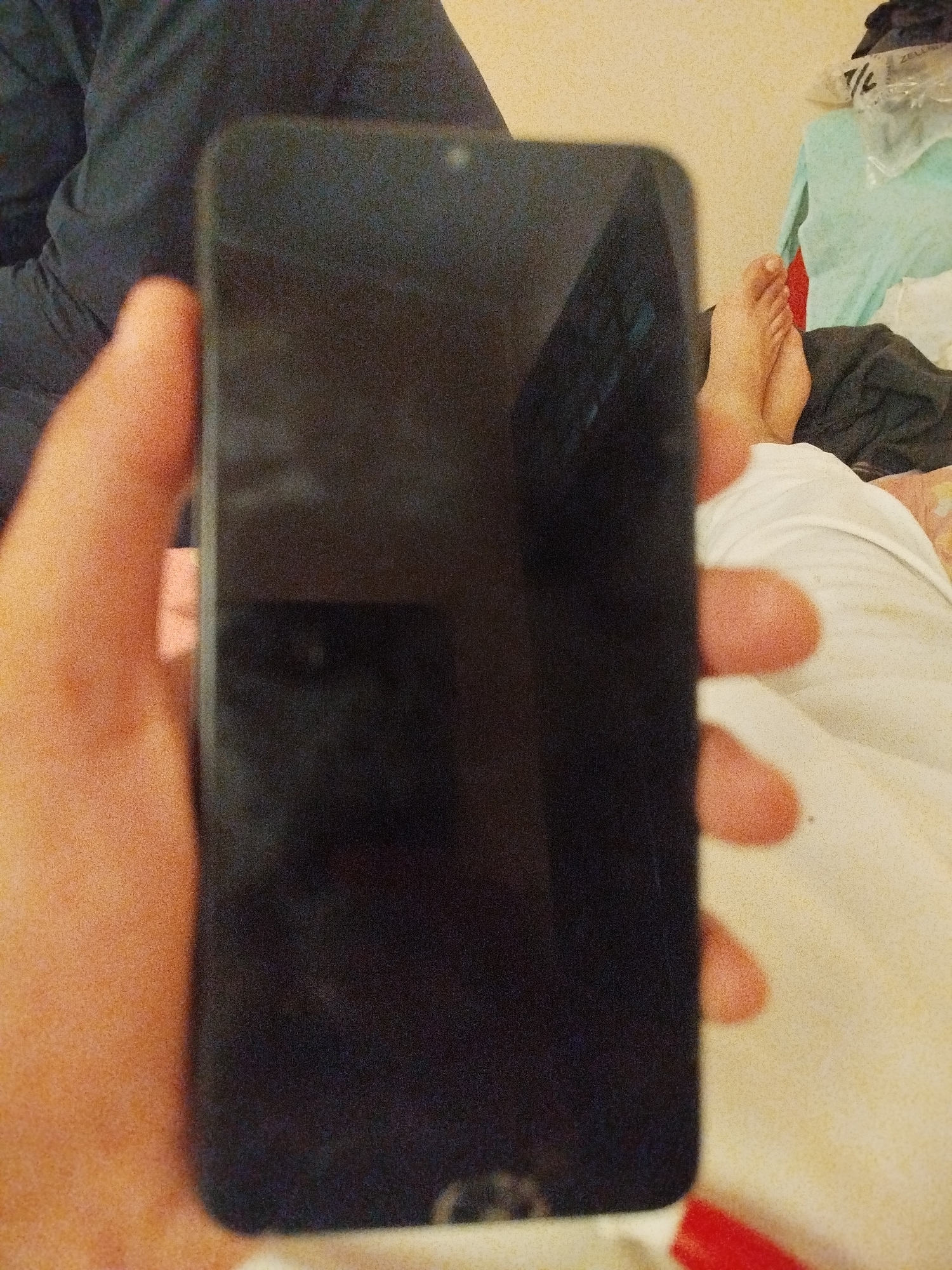 A person's hand is holding a black smartphone in a horizontal orientation. The phone's screen is black and reflects some of the surroundings. In the background, a person's feet are visible, as well as some clothing and a wall. There is no indication of the location being in Lassan, Pakistan; this is a domestic scene.