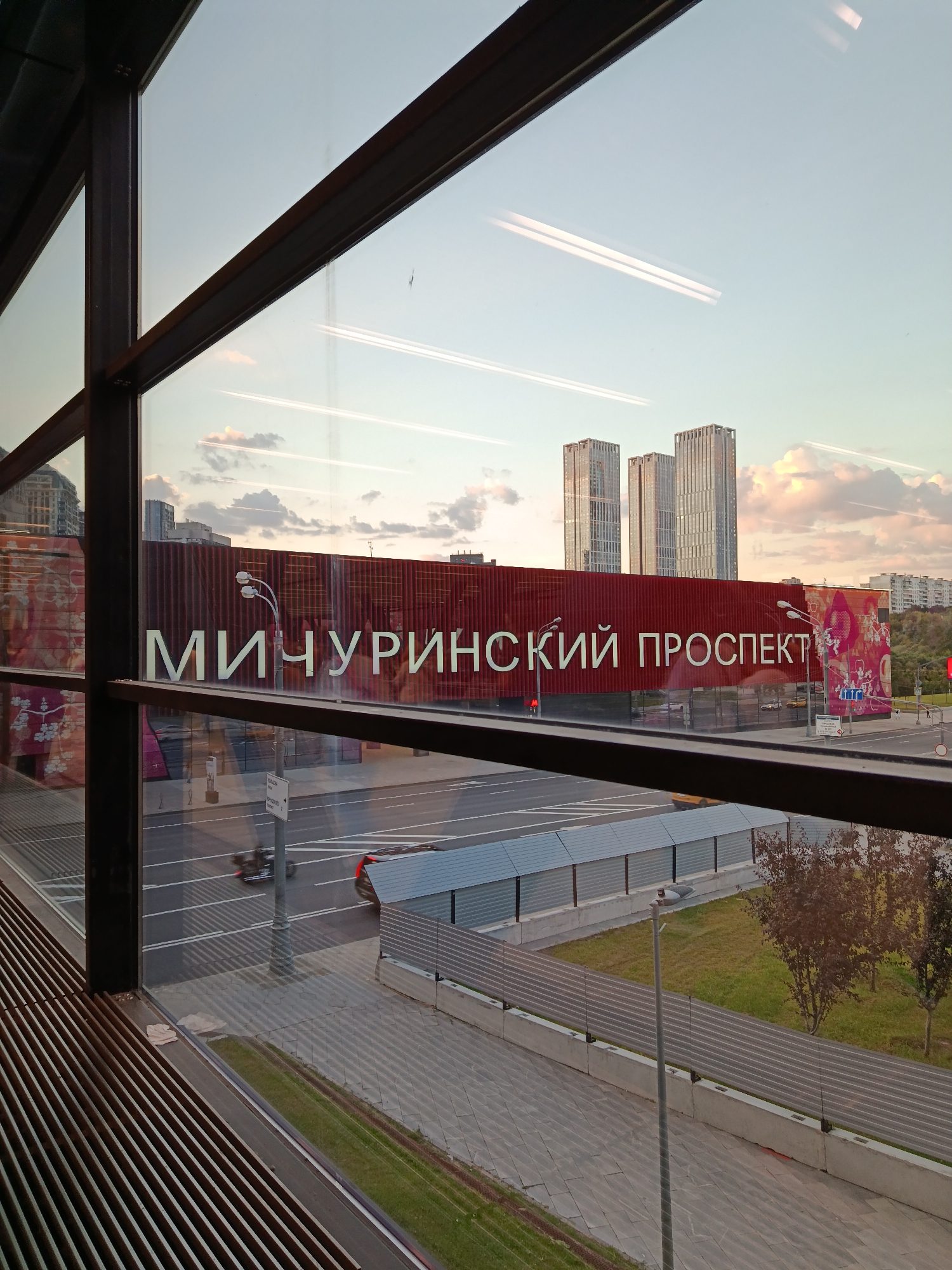 A wide-angle, exterior shot captures a view through a large window, looking out at a city landscape at dusk. The window frame, made of dark metal beams, is visible in the foreground. The sky is a pale blue with scattered clouds and streaks of light, possibly from contrails.
Dominating the scene is a large, dark red building with the white Cyrillic text "МИЧУРИНСКИЙ ПРОСПЕКТ" prominently displayed across its facade. To the right of this building, a smaller section features a graphic design in pink and white. Behind the red building, three tall, modern skyscrapers rise into the sky, their windows reflecting the evening light.
Below the building, a street with multiple lanes of traffic is visible. A car and a motorcycle are captured in motion, with a red car parked to the far right. A pedestrian walkway with patterned paving stones runs along the bottom of the frame, bordered by grass and trees. A street lamp stands on the walkway, and a metal barrier with a corrugated roof extends across the middle ground. The overall impression is one of a modern urban environment at the end of the day.