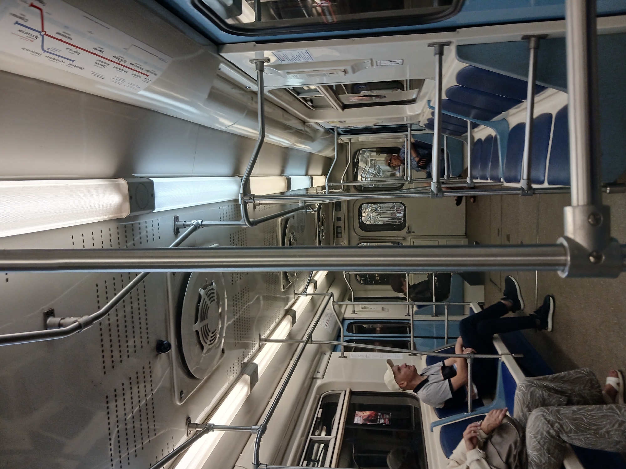 The image depicts the interior of a passenger train car, likely a metro car, in Nizhny Novgorod, Russia. The car has a utilitarian design with blue and white seating arranged along the sides. Several passengers are visible: a man in a white cap and a grey and black striped shirt is seated on the left, facing forward with his arms crossed. Further down the car, another passenger is partially visible. The ceiling features fluorescent lighting and ventilation grilles. A detailed metro map, indicating lines and stations, is displayed on the right wall. The overall environment is that of public transportation, functional and sparsely decorated, typical of urban rail systems.