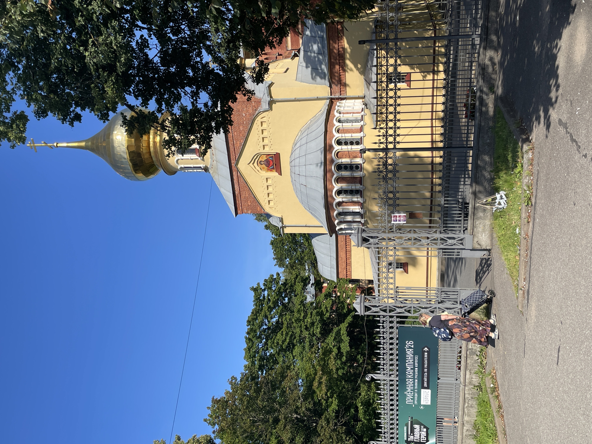 In Saint Petersburg, Russia, a woman with a backpack and a rolling cart walks away from the entrance of a yellow-colored church with a golden dome. The church has a red brick facade and is surrounded by a black metal fence. A banner with Russian text is visible on the fence, advertising an admission campaign for 2026. Trees with green foliage are present on the left and right sides of the image, and the sky is clear and blue. A power line is visible against the sky.