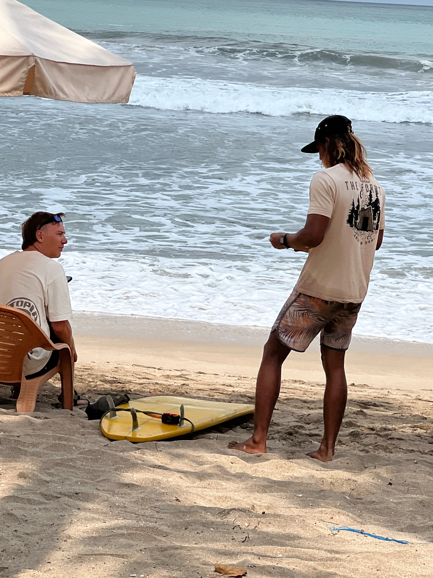 Two men are on a sandy beach in Kuta, Indonesia, with the ocean and waves in the background. The man on the left is seated on a brown plastic chair, wearing a white t-shirt and sunglasses on his head. The man on the right is standing, wearing a light-colored t-shirt with a graphic print and patterned swim shorts. He is facing the ocean and appears to be holding something in his hands. A yellow surfboard is lying on the sand in front of him. An umbrella is partially visible on the left side of the image. The water is choppy with white foam from the waves.