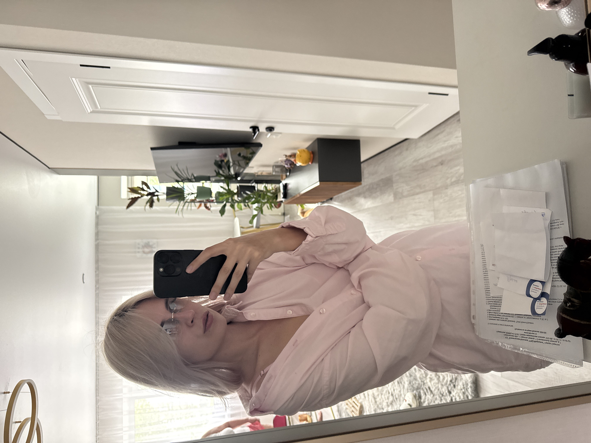 A selfie shows a blonde woman with glasses holding a black cell phone and wearing a pink button-down shirt in a bathroom. In the background is a counter with a TV and plants, a white cabinet, and a tiled floor. Papers and objects are visible in the mirror's reflection. The location cannot be definitively determined as Saratov, Russia based on the image.