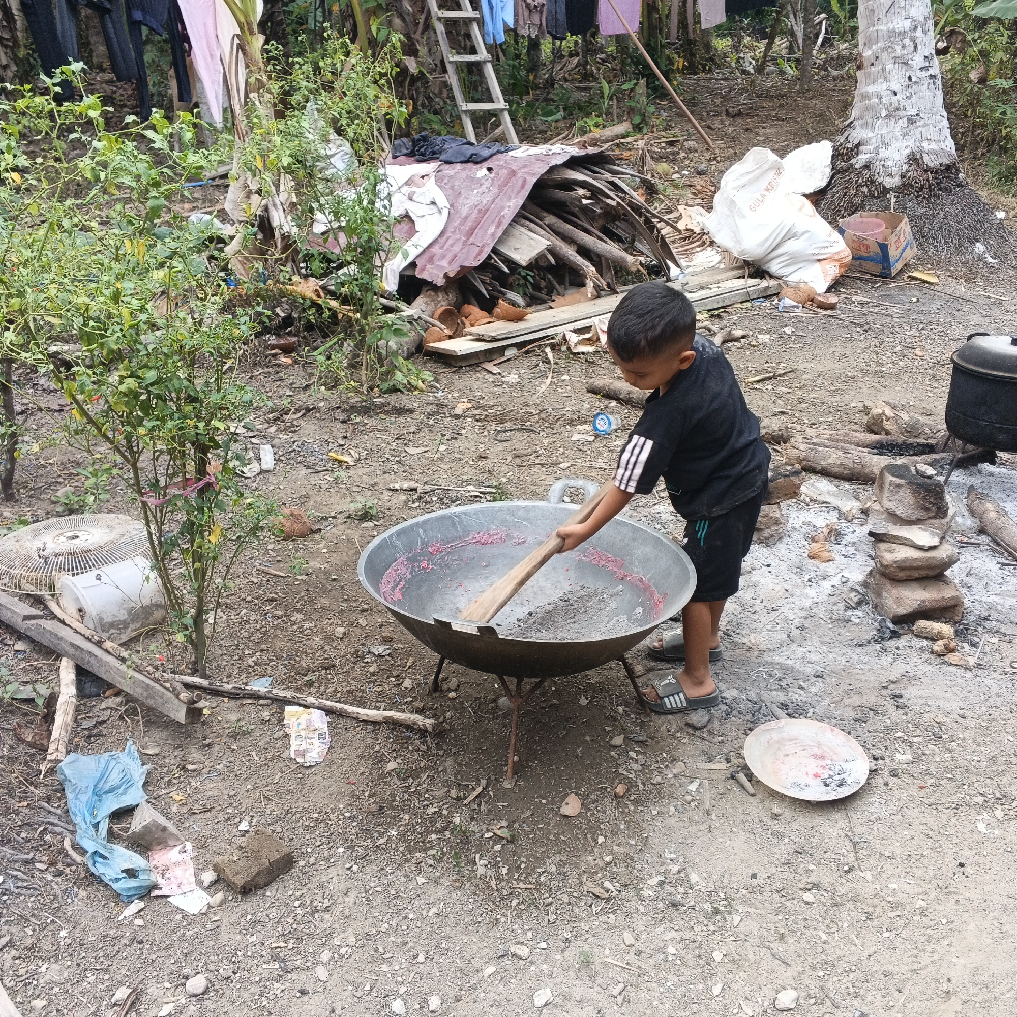The image depicts an outdoor scene in an informal setting, possibly a yard or backyard in an urban or rural area. The ground is composed of dirt and gravel, with scattered debris including pieces of wood, plastic bags, and what appear to be coconut shells. In the foreground, a young boy wearing a black t-shirt with white stripes on the sleeves and black shorts is standing and stirring a large, round, metal wok with a wooden spoon. The wok contains a reddish substance and some ash. To the right of the boy, a small stack of stones is visible, and beyond that, a large pot is sitting on a makeshift stove. Further in the background, a pile of firewood covered with a reddish corrugated metal sheet is present. A wooden ladder leans against a tree. Laundry, including a pink garment and dark clothing, is hanging on a line. A white plastic fan and a white bucket are also visible near some green plants. A large tree with a textured trunk is located on the right side of the image. The overall impression is one of everyday life in a place where resources may be limited.