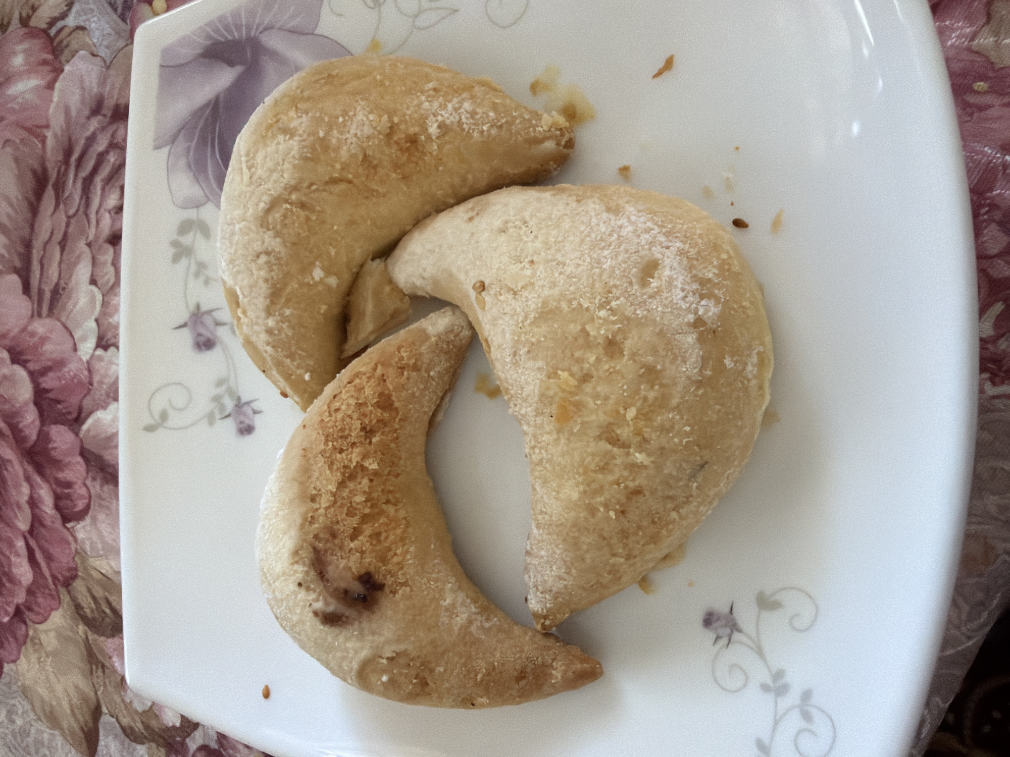Three crescent-shaped pastries dusted with powdered sugar are arranged in a triangular formation on a white plate. The plate has a floral pattern with purple and green accents. The pastries appear to be baked goods, possibly cookies or small cakes, with a slightly golden-brown exterior and some darker browning on one side of the top pastry, suggesting it may have been slightly overcooked or contained a filling that browned. The background is a patterned tablecloth with large pink and purple floral motifs. The scene is lit from above, casting subtle shadows. There are no people or animals visible. The image is taken from a top-down perspective.