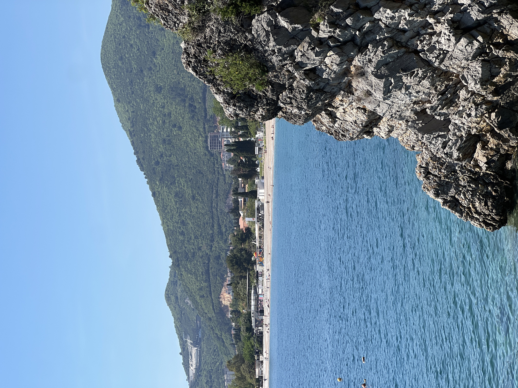 This image captures a vibrant coastal landscape in Budva, Montenegro, viewed from an elevated perspective on a bright, clear day.
In the foreground, a rugged, dark grey rock formation, partially adorned with green shrubs, descends steeply from the bottom right, framing the scene. Beyond this rocky outcrop, the calm, deep blue waters of the Adriatic Sea stretch across the mid-ground, showing subtle ripples and a few scattered yellow and red buoys. Along the coastline, a long, light-colored beach is visible, populated by numerous small figures of people. They appear to be relaxing on sun loungers, walking along the shore, or wading in the shallow sea, indicating typical beach activities.
Behind the beach, a strip of modern white and light-toned buildings, including multi-story structures, line the coast, interspersed with green trees and vegetation. Further inland and dominating the left side of the image, a large, lush green mountain rises, densely covered in trees, extending high into the clear, light blue sky. Hints of distant infrastructure or buildings are visible on the mountain slopes in the background.
The time of day appears to be mid-morning or early afternoon, with bright sunlight illuminating the entire scene and casting distinct shadows on the rocky cliff. The weather is perfectly clear and sunny, creating an inviting atmosphere for beachgoers. The overall impression is that of a popular and picturesque resort town during peak season. No discernible text is visible within the image.