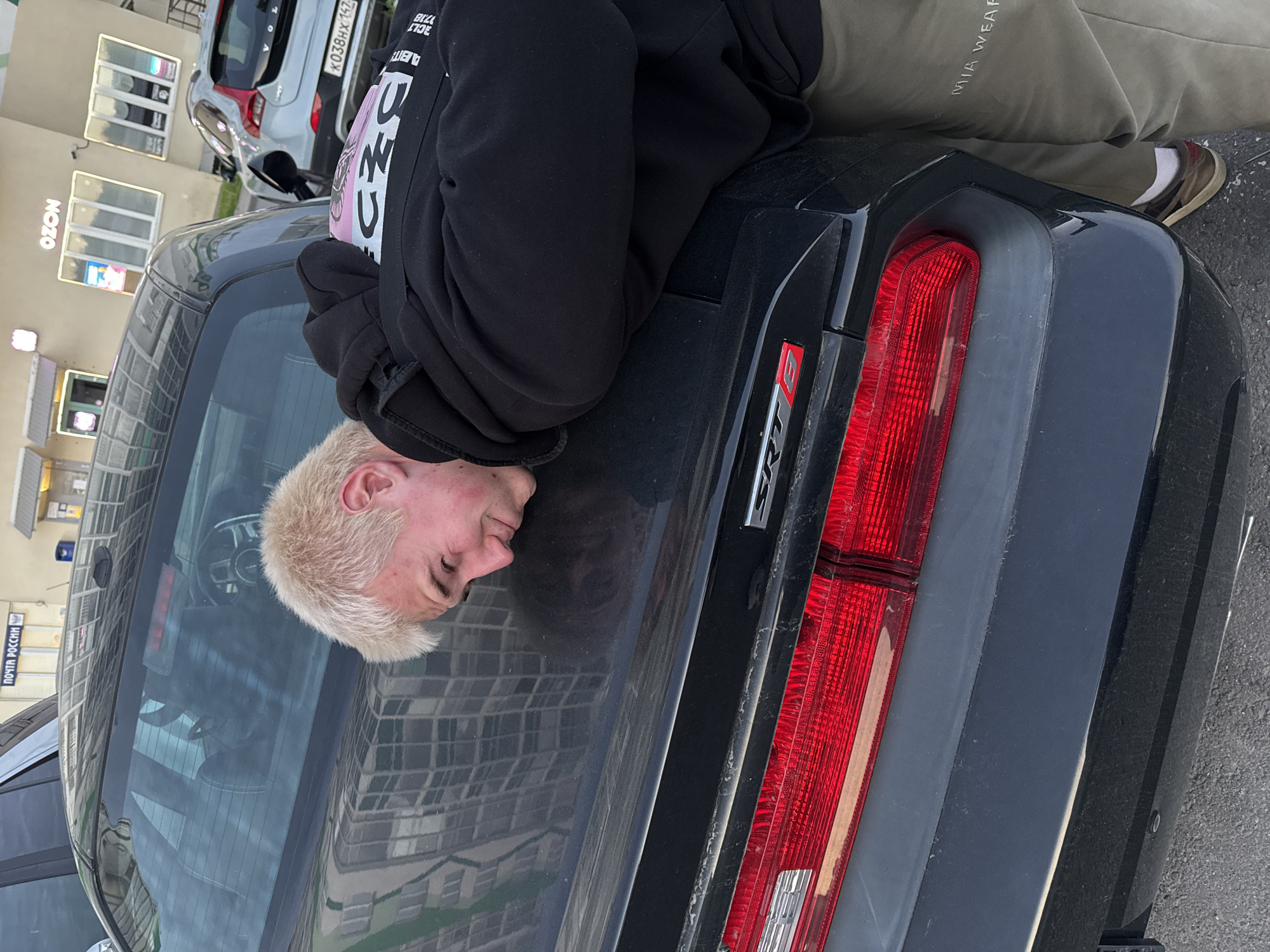An image, rotated on its side, displays a young person with short, bleached blonde hair lying face down on the trunk of a black Dodge Challenger. The person is wearing a black hooded sweatshirt, olive green pants with "MIA WEAR" written on them, and brown sneakers with white socks. Their face is pressed against the car's reflective surface, showing a distorted reflection of their features and surrounding buildings. The black car features prominent red rectangular taillights and an "SRT" emblem on its rear.
In the background, an urban setting is visible with buildings. One building prominently displays "OZON" signage, and another has "ПОЧТА РОССИИ" (Russian Post) text. A silver/grey Volvo SUV is parked behind the black car, bearing a Russian license plate "K038HX 147." The scene is brightly lit, indicating daytime. The person's pose appears deliberate, with a calm or contemplative expression. The car's bumper area shows some dust accumulation. Additional partial text, "3CL" and "17718", is faintly visible on the person's hoodie.