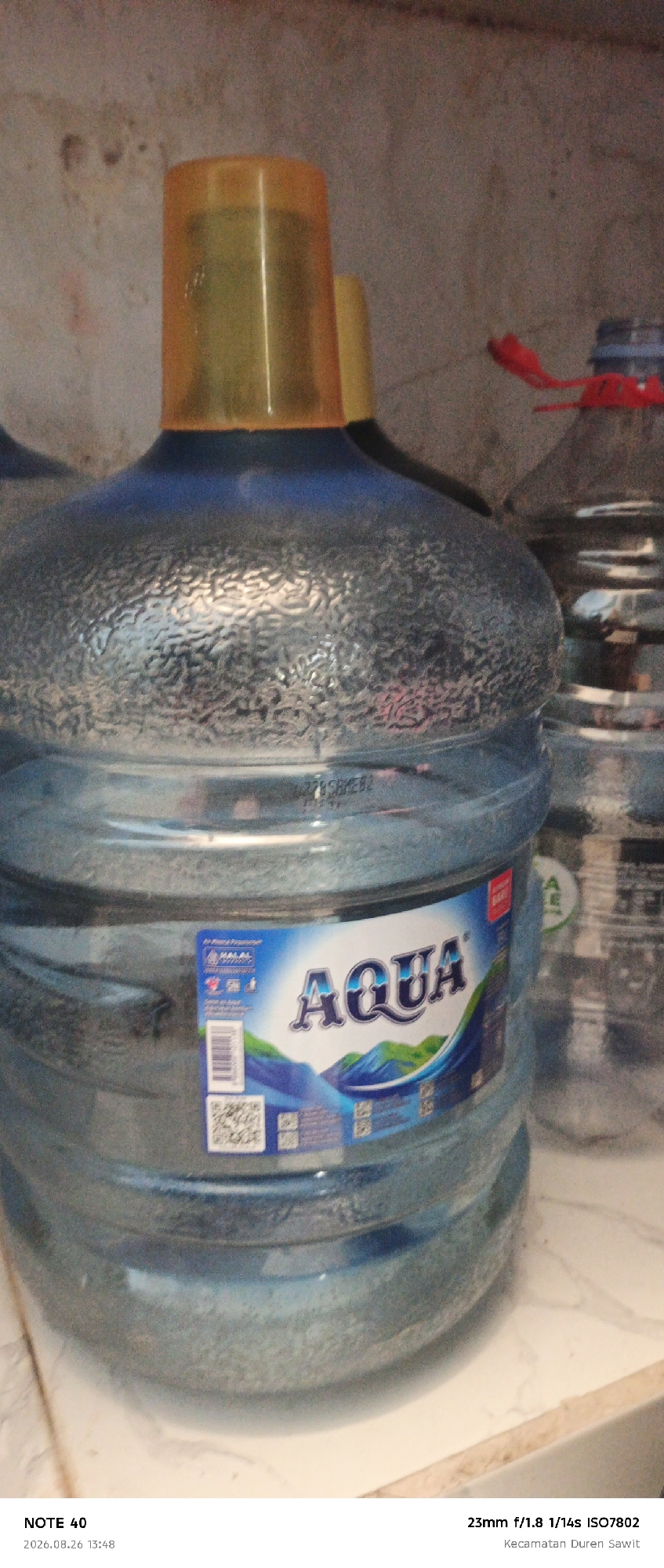 A close-up, eye-level, medium shot captures two large, transparent plastic water jugs against a textured, beige wall. The jug in the foreground is the primary subject, its entire body visible. It has a textured, bumpy surface and contains clear liquid. A prominent blue label on its front displays the white "AQUA" logo with a mountain graphic below. To the left of the logo is a smaller label with text and a QR code. The jug's cap is a bright yellow plastic. Behind this jug, a second similar jug is partially visible, featuring a red cap with a pull tab. The background wall shows signs of wear and discoloration. The lighting is dim, casting soft shadows and highlighting the textured surfaces of the jugs. The lower right corner of the image displays text that includes "NOTE 40" and a date and time stamp, followed by camera settings.