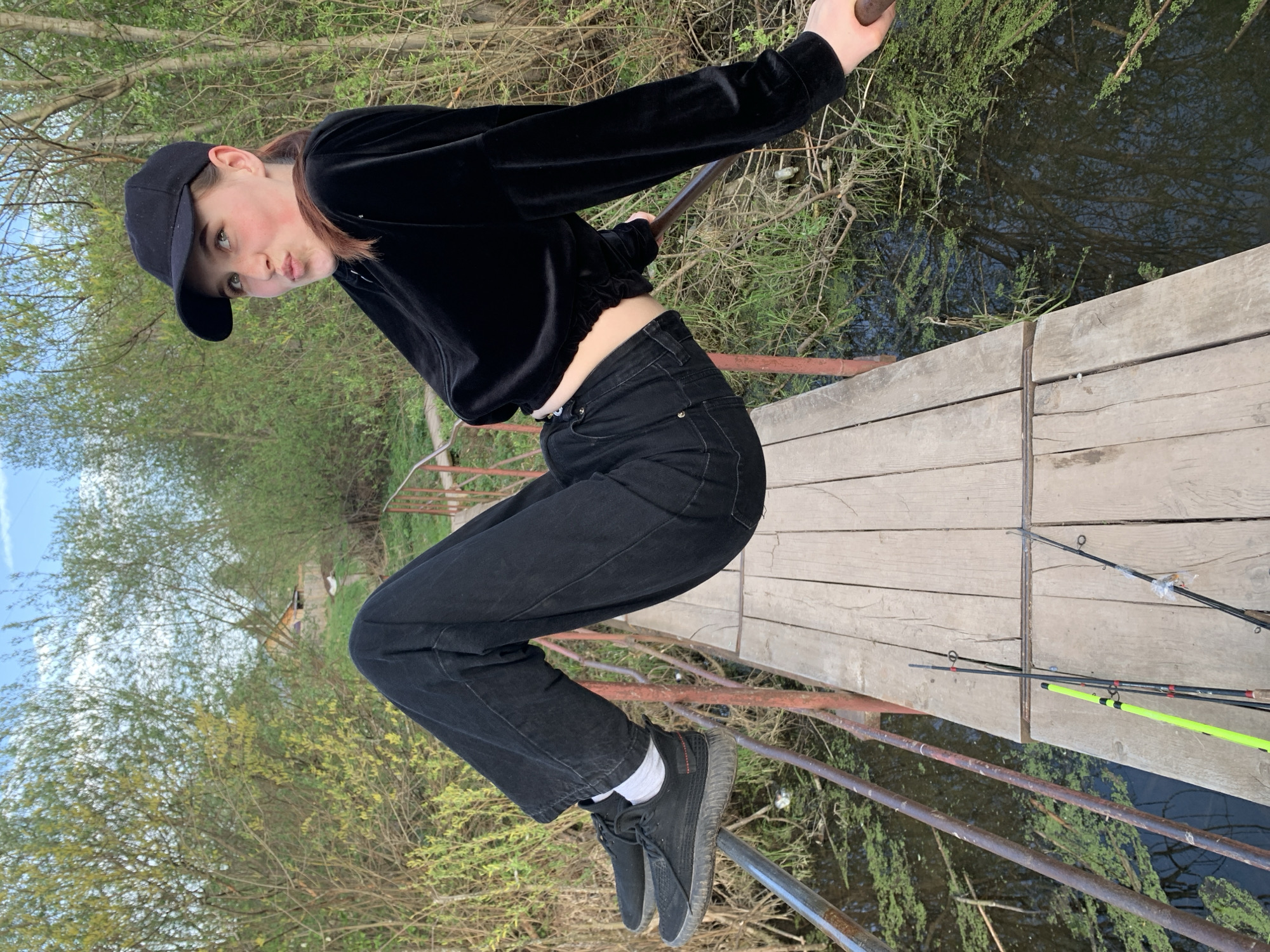 A young woman stands on a wooden bridge over water. She wears a black cap, a black velvet crop top, black jeans, and black sneakers with white socks. She leans forward, holding onto a railing with one hand while looking at the camera with a pursed-lip expression. Fishing poles lean against the bridge. The environment appears to be a natural area, with trees visible in the background. The scene is in Фоки, Russia.