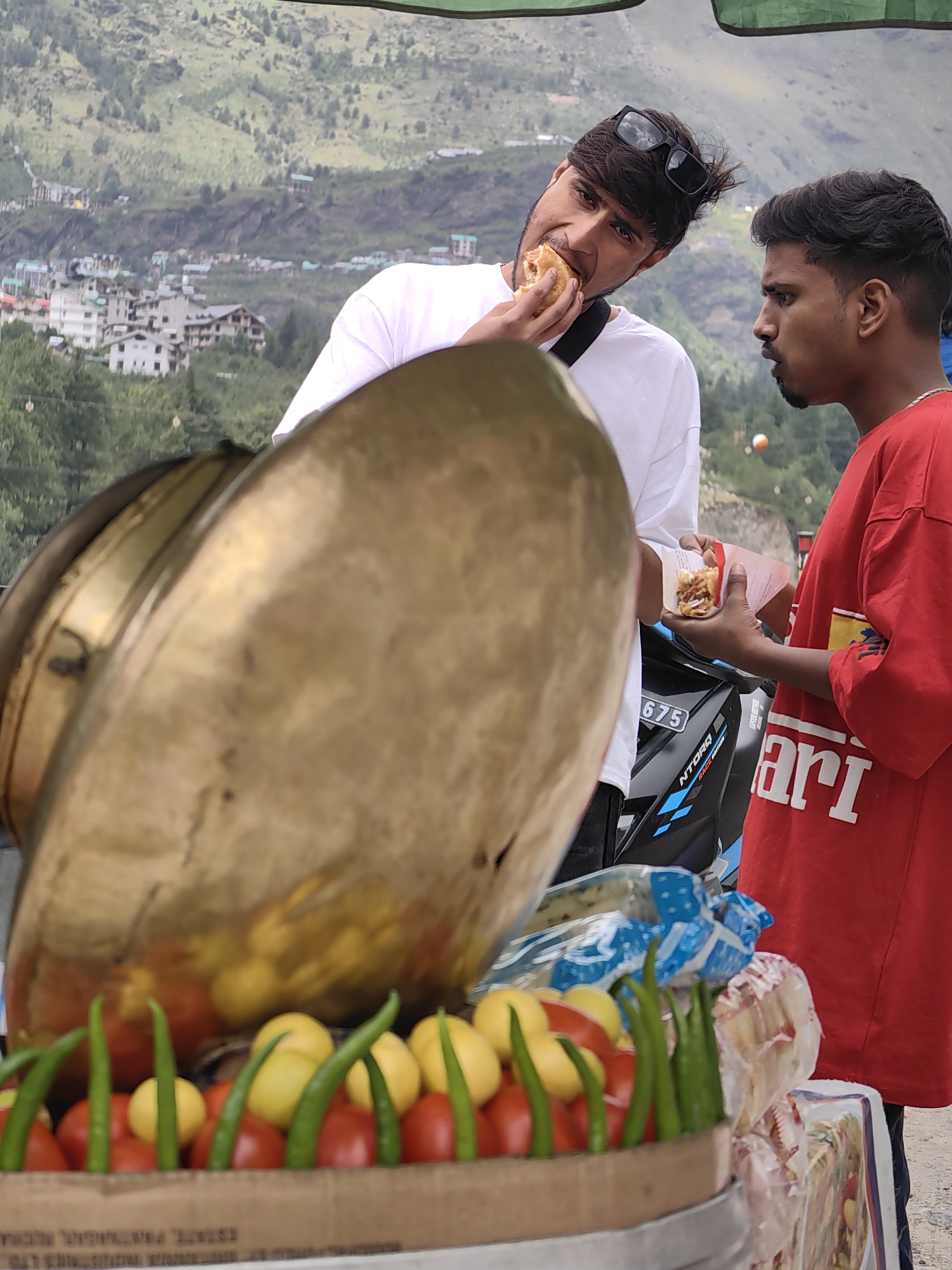 Two young men are standing outdoors with a blurred mountain village in the background. On the left, a man in a white t-shirt is taking a bite of a sandwich. He is wearing sunglasses on his head. On the right, a man in a red t-shirt is holding a sandwich in his hand and appears to be speaking to the other man. In the foreground, there is a display of fresh produce, including tomatoes and green chilies, contained in a cardboard box. There is also a large, golden, bowl-shaped object in the foreground that partially obscures the view of the two men. A motorcycle is parked behind the men. The overall scene suggests a street food vendor setting.