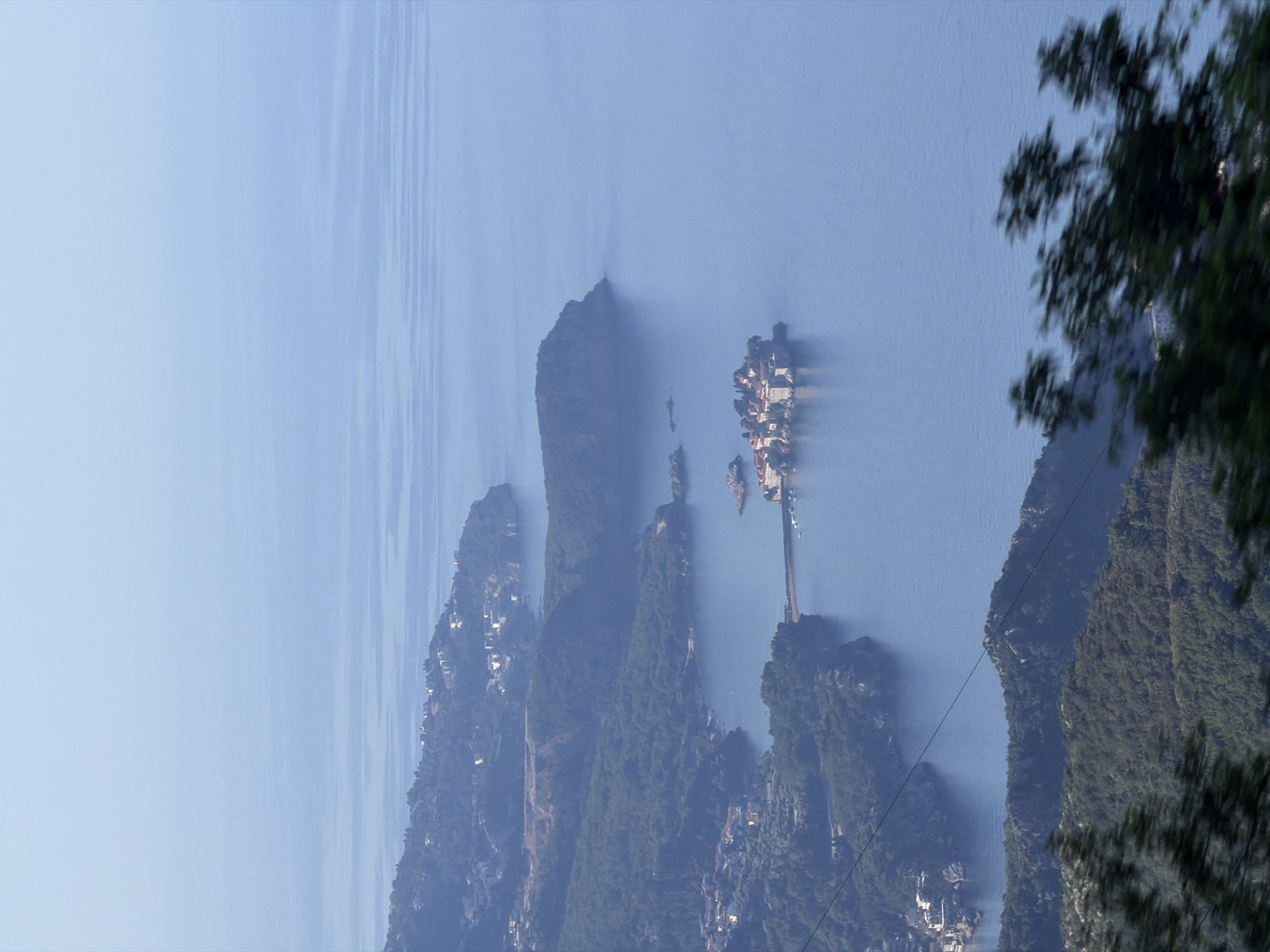 An aerial, slightly tilted, full shot captures the coastline and a small island settlement in Stanišići, Montenegro. The sky is a hazy, light blue, fading into a slightly darker blue towards the horizon, where it meets the sea. The sea itself is a calm, pale blue, with subtle horizontal lines indicating gentle ripples.
The coastline is characterized by steep, densely forested hills that descend sharply to the water. Numerous white buildings, indicative of a settlement, are scattered along the hillsides, some appearing to be villas or homes. A thin, dark line, possibly a cable or power line, stretches diagonally across the middle ground, obscuring some of the view of the landscape.
In the middle of the sea, connected to the mainland by a thin causeway, sits a small, picturesque island settlement. The island is covered with buildings featuring red-tiled roofs, and appears to be fortified with stone walls. Several small boats are visible near the causeway.
In the foreground, out-of-focus dark green foliage, possibly tree branches, blurs the bottom of the image, creating a sense of depth and suggesting the viewer is looking from a higher vantage point. The overall atmosphere is serene and picturesque, with the natural landscape dominating the scene.