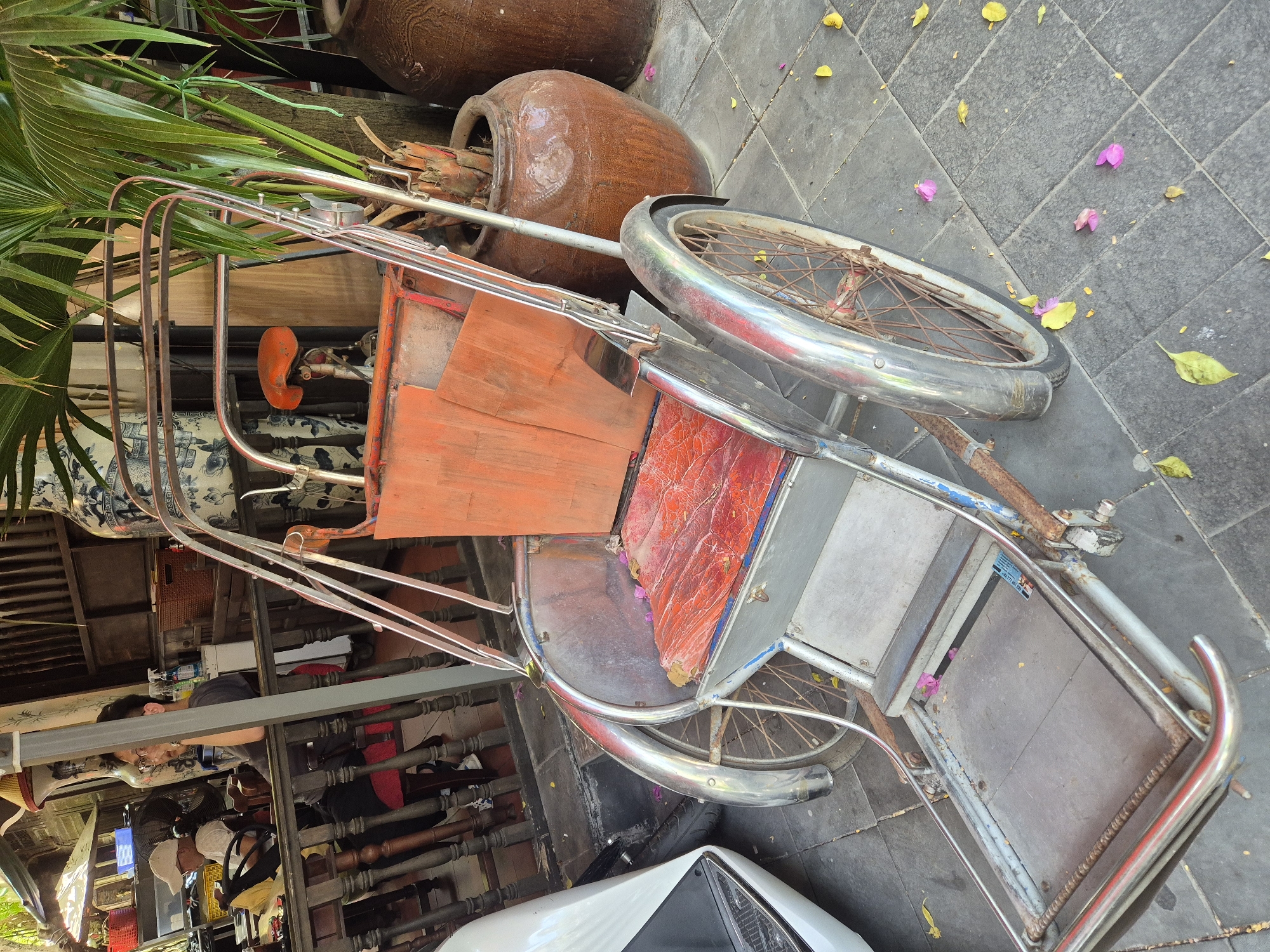The image displays a pedicab parked on a tiled sidewalk in Da Nang, Vietnam. The pedicab is made of metal with a red seat and orange wooden panels. It has three wheels, with the front wheel being larger than the rear ones. A white scooter is partially visible to the left of the pedicab. In the background, a railing separates the sidewalk from a seating area with wooden tables and chairs. Two people are seated at one of the tables, one wearing a white cap and the other a dark shirt and glasses. Large ceramic pots and a potted plant are situated behind the pedicab. Fallen yellow and pink petals are scattered on the ground.