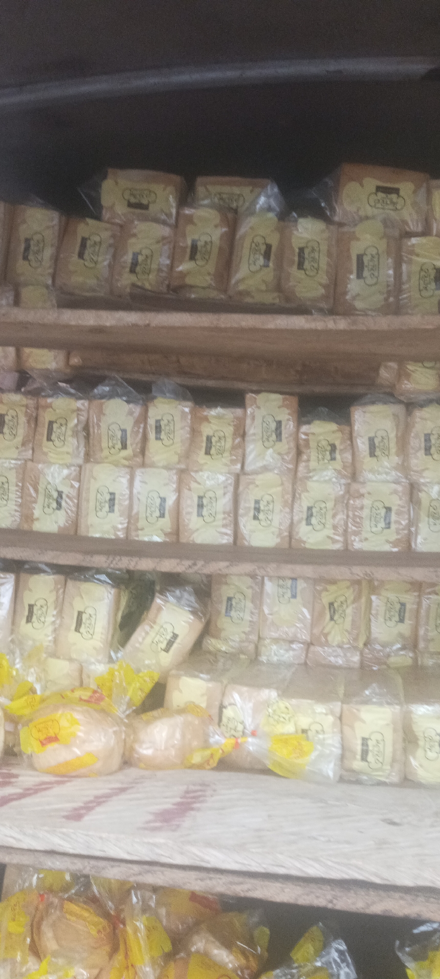 Image shows several shelves of packaged bread loaves in Kuru Dungus, Nigeria. Each loaf is individually wrapped in clear plastic with a yellow label. The loaves are densely packed on the shelves. The shelves themselves are wooden. The overall lighting in the image suggests an indoor or sheltered environment.