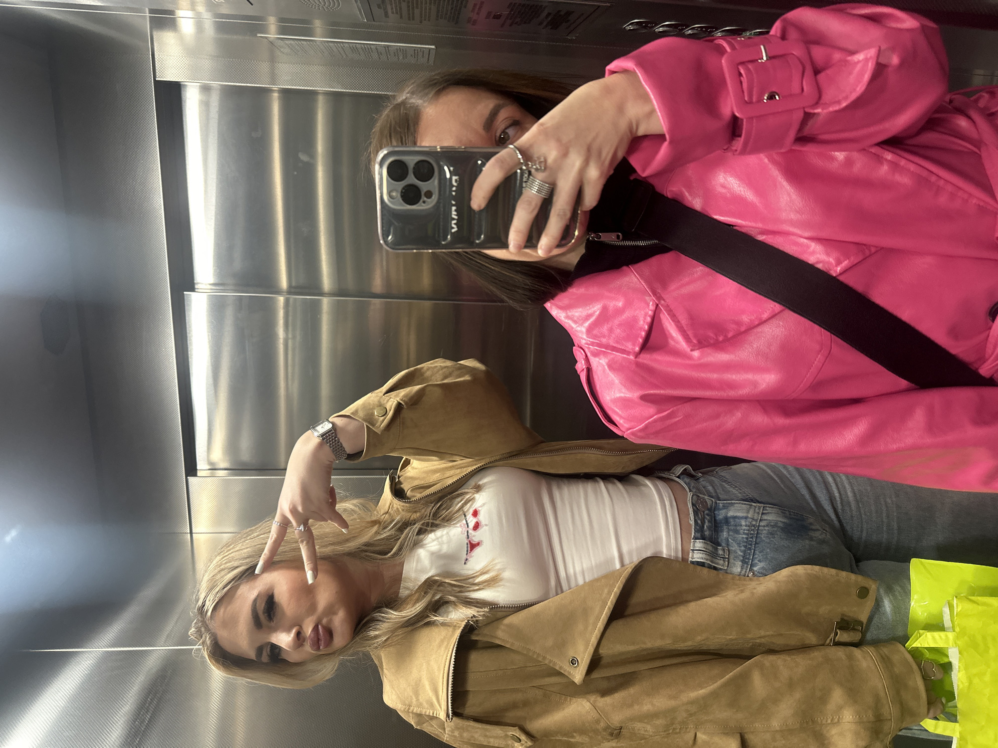 Two young women are depicted taking a selfie inside an elevator with brushed metallic walls in Kazan, Russia.
The woman on the left, with blonde hair styled in waves and visible makeup, poses with her lips pursed and her right hand displaying a "peace" sign. She wears a tan/light brown suede-like jacket over a white fitted crop top featuring a small red graphic. Light-wash blue jeans and a silver-toned rectangular wristwatch on her left wrist are also visible. A neon green shopping bag is partially visible in her left hand at the bottom left.
The woman on the right is largely obscured by the smartphone she holds, with only her right eye and forehead visible. She has dark, shoulder-length hair. She wears a bright fuchsia/hot pink trench coat, possibly made of a shiny faux leather material, with a prominent buckled cuff on her right sleeve. A black cross-body bag strap crosses her chest, and a black top is visible underneath her coat. She holds a smartphone, appearing to be an iPhone Pro Max model, encased in a clear or translucent grey case with a visible "DEBOSS" logo. Multiple rings are present on the fingers of her right hand, including a chunky silver-toned ring and another with a small charm.
The elevator interior features metallic, possibly stainless steel, wall panels with a subtle textured pattern. Two vertical elevator doors are visible behind the subjects, reflecting the interior lighting.