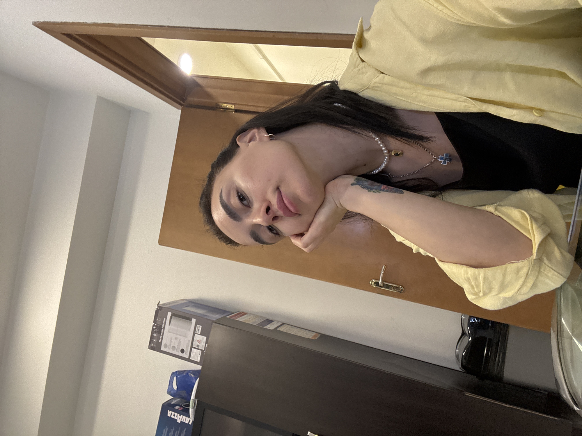 A person with dark hair is resting their face on their hand, facing the camera. They are wearing a black top underneath a yellow button-down shirt. A pearl necklace and a silver necklace with a cross pendant are visible. The background includes a brown door frame and a light-colored wall. Below, there is a dark-colored cabinet with a box of "Lavazza" coffee and another box. A dark, cylindrical object (possibly a coffee grinder) sits on top of the cabinet, near a metallic object. The specific location in Tbilisi, Georgia, cannot be determined from the image.