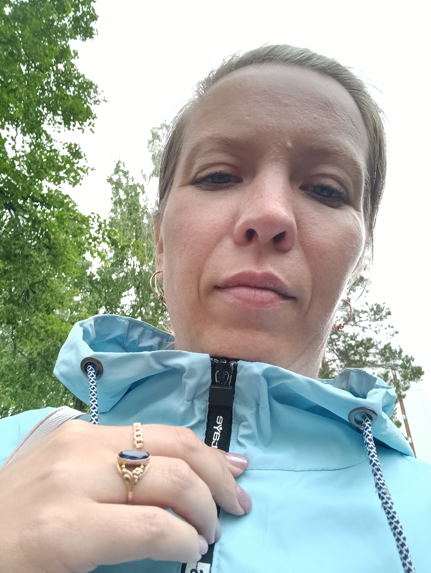 A person, presumably female, is taking a selfie outdoors in Chaikovsky, Russia. The person is fair-skinned with light hair pulled back. They are wearing gold hoop earrings and a gold ring with a dark oval stone. The person is wearing a light blue jacket with a hood that is partially up. The zipper of the jacket is visible. Trees are visible in the background, suggesting the location may be a park or a green area within the city. The sky appears overcast.