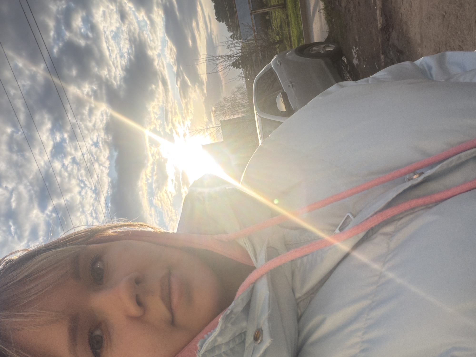 The image is a selfie captured outdoors, rotated 90 degrees clockwise. In the foreground, a young person is visible, looking directly at the camera. They have light-colored hair, and their eyes are a prominent feature. They are wearing a light blue or grey puffer jacket with visible pink drawstrings around the collar, and a pink garment underneath.
The background features a dramatic sky filled with scattered clouds, ranging from white to darker grey. The sun is low in the sky, positioned behind and to the right of the person, casting a bright lens flare across the upper and middle portions of the image, indicating either sunrise or sunset. The intense sunlight creates a warm glow.
Further back, to the left of the person, a light-colored car (possibly white or silver) is parked on what appears to be a dirt or gravel roadside. Beyond the car, there are trees and sparse vegetation, suggesting a rural or suburban setting. Power lines are visible stretching across the upper left quadrant of the sky.
The overall mood is bright and somewhat serene, characterized by the natural light and expansive sky. There is no visible text in the image.