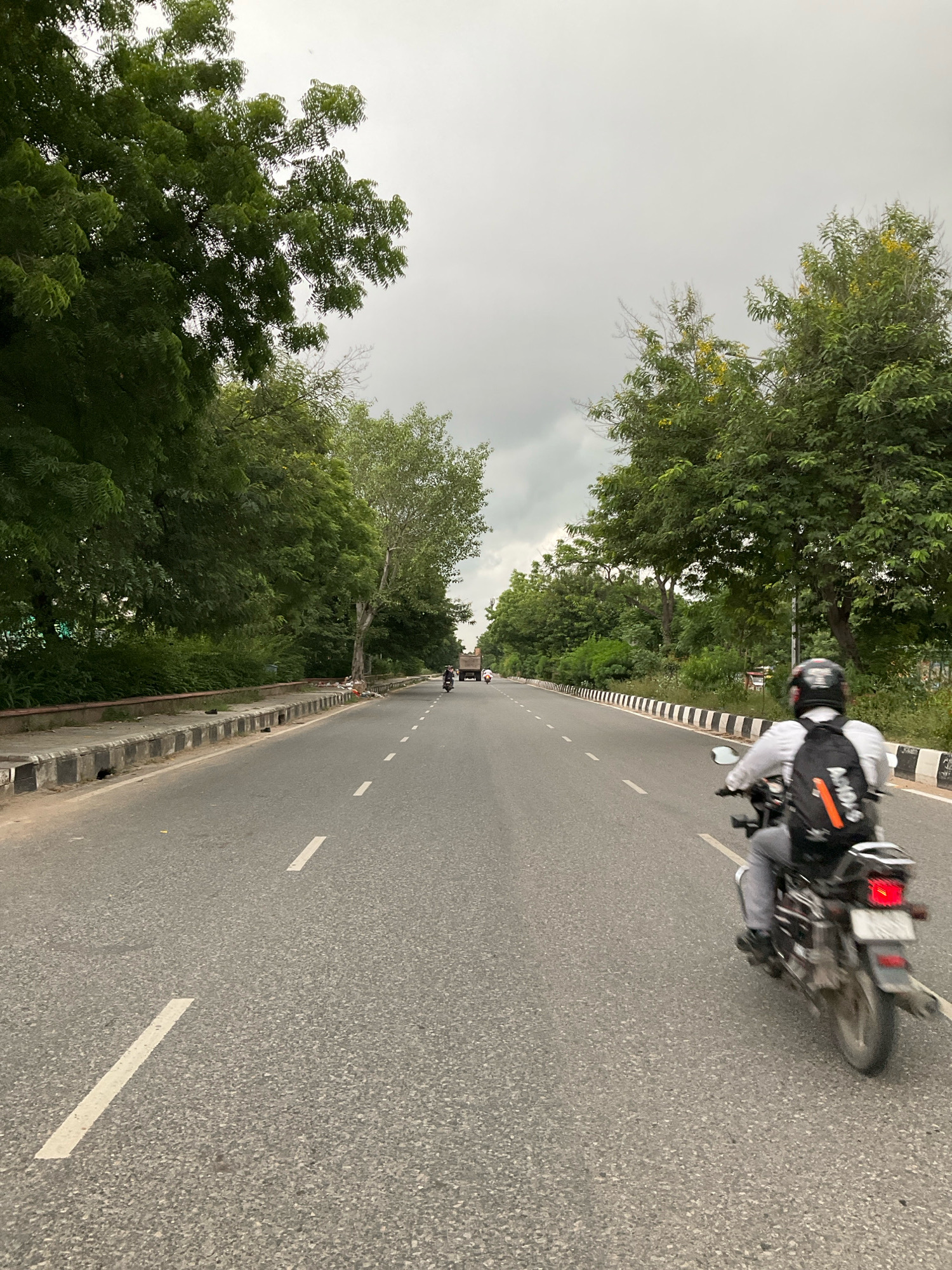 A motorcycle rider with a black helmet and backpack is traveling on a two-lane road lined with trees and greenery. The road has white dashed lane markers. In the distance, there are other vehicles and more trees. The sky is overcast, suggesting it might be a cloudy day. The location appears to be within the Jaipur Municipal Corporation, India, likely on a route with significant vegetation alongside the roadway. The overall scene depicts everyday traffic on a road surrounded by nature.