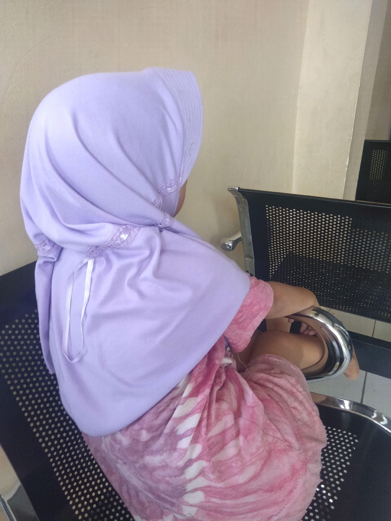 A person wearing a light purple hijab and a pink tie-dye dress is seated on a black chair. The chair has a perforated metal back and a chrome armrest. The person's back is to the camera, and they are looking to the right. The background is a beige wall. The scene appears to be indoors, possibly in a waiting area.