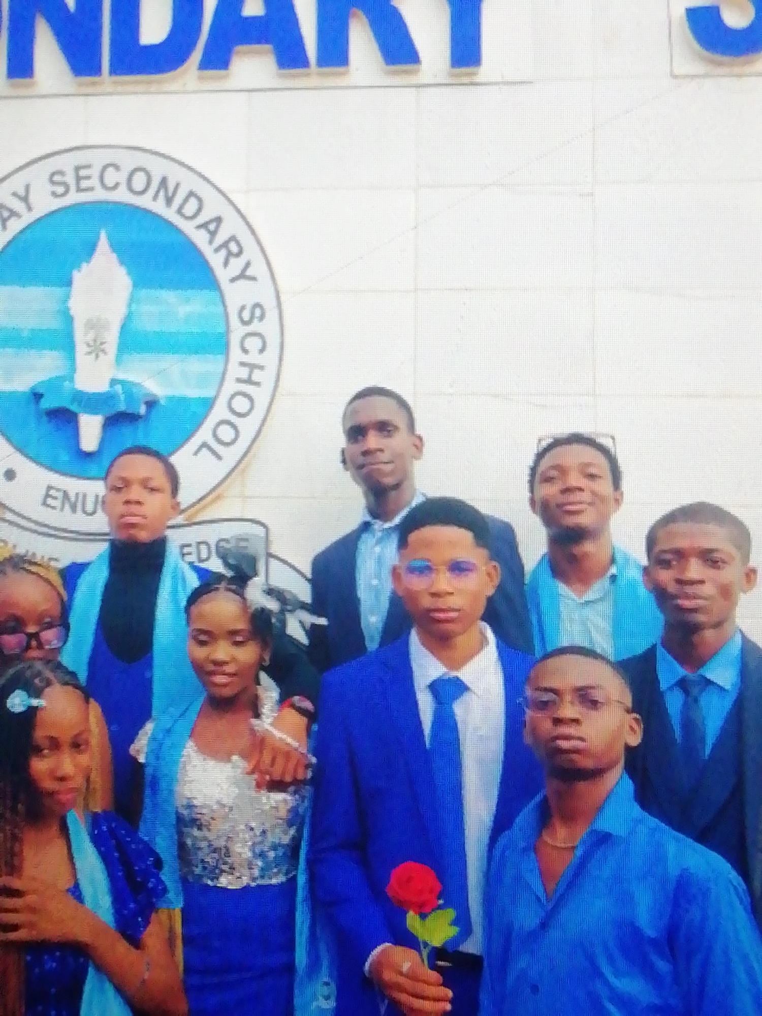A group of eight young people, likely students, are captured in a posed photograph against a light-colored wall. The group consists of five males and three females, predominantly dressed in various shades of blue and white, with some black and silver accents, suggesting formal attire for a school event. Many of the males wear blue blazers, white shirts, and blue ties, while the females are in blue dresses or skirts, one featuring a sparkly silver top. A male in the foreground holds a single red rose.
In the background, a large circular emblem is prominently displayed on the wall. The text "GATEWAY SECONDARY SCHOOL" is visible around its circumference, with "ENU" (likely an abbreviation for Enugu) inside the circle, and the partial word "EDG" below it, possibly part of a motto. Above the emblem, large blue letters spell out "NDARY S," confirming the school context. The emblem's central design features a stylized monument or torch with a star-like symbol at its base.
The individuals exhibit neutral to slightly smiling expressions for the camera. The bright lighting suggests an indoor setting or a brightly lit outdoor area. The visible school name and formal attire strongly indicate a school-related event, such as a graduation or valedictory ceremony, taking place in Enugu, Nigeria.