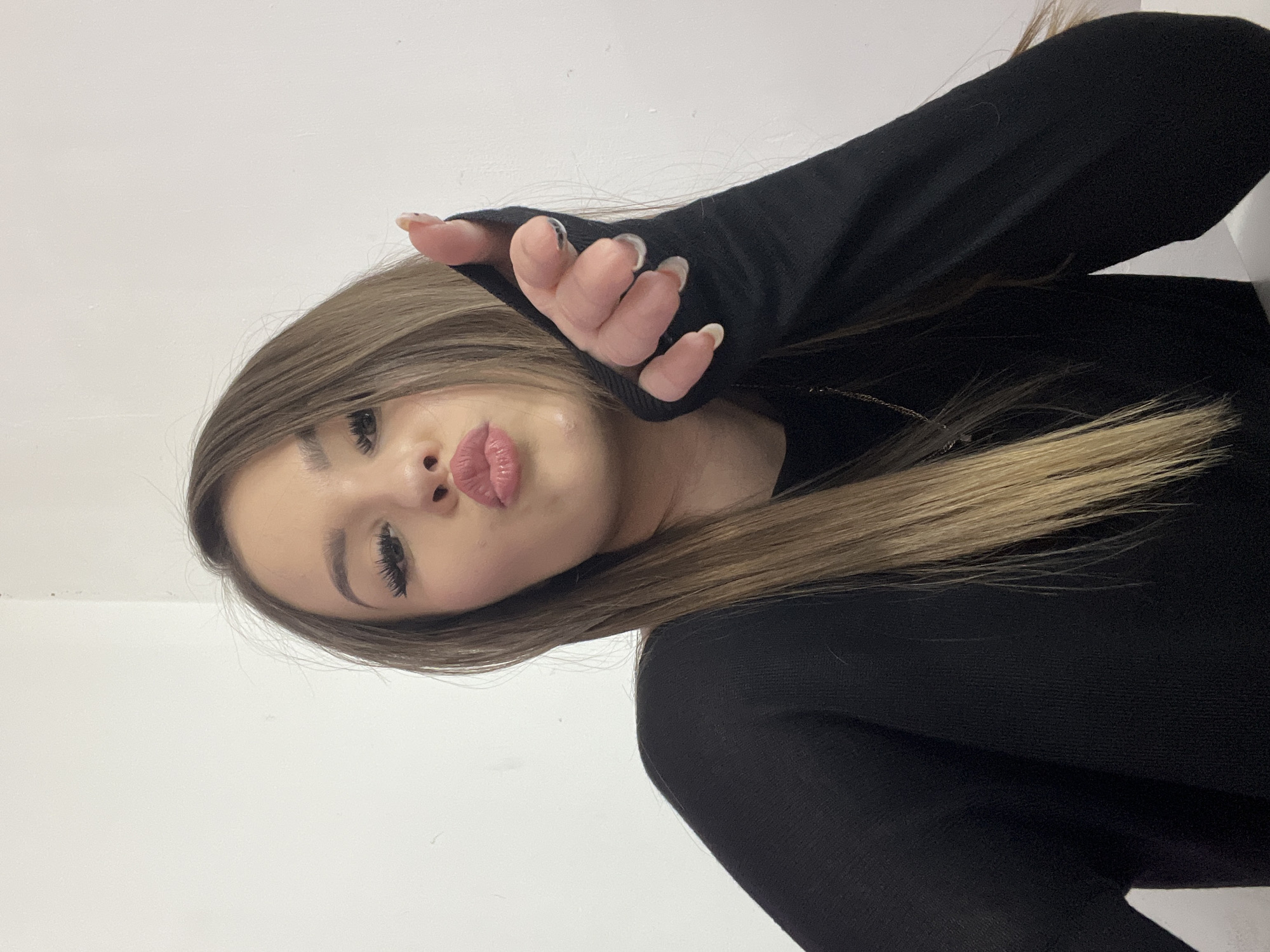 The image shows a young woman with long, blonde-streaked brown hair, wearing a black top with thumbholes. She is leaning forward and puckering her lips, with her right hand extended towards the camera, pointing. Her nails are painted with a clear or light pink polish. The background is a plain white wall. There is no discernible indication of the location being in Noviy, Russia, or any other city. The scene is a close-up portrait of the woman.