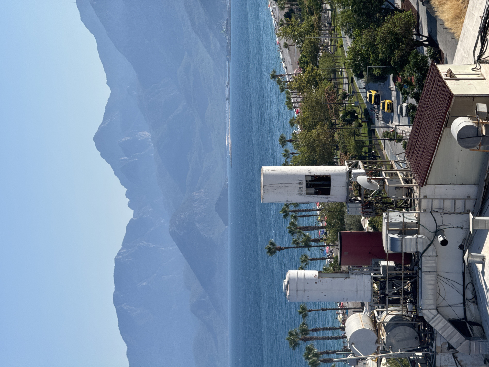 This image depicts a coastal scene in Konyaaltı, Turkey. The foreground is dominated by the rooftops of industrial structures, possibly communication towers, cluttered with various equipment, pipes, and cables. These structures are primarily white and grey with some red accents.
Beyond the rooftops, a tranquil blue sea stretches towards a distant coastline. Along the shore, a beach lined with palm trees is visible, with what appears to be beach umbrellas and people enjoying the seaside. Further inland, a road runs parallel to the coast, with several yellow vehicles, possibly taxis, driving on it. Lush green trees flank the road, creating a vibrant contrast with the blue of the sea and sky.
In the background, a majestic mountain range rises, its slopes shrouded in a soft haze, suggesting either distance or a particular atmospheric condition. The sky is a clear, pale blue, indicating a bright, likely sunny day.
The image captures a moment of everyday life in a coastal resort town, blending urban infrastructure with natural beauty. There is no discernible activity or event taking place beyond the general scene of a populated beach and a functioning roadside. The overall impression is one of a sunny day in a Mediterranean setting. Visible text on one of the structures reads "GÜN İŞİ".
