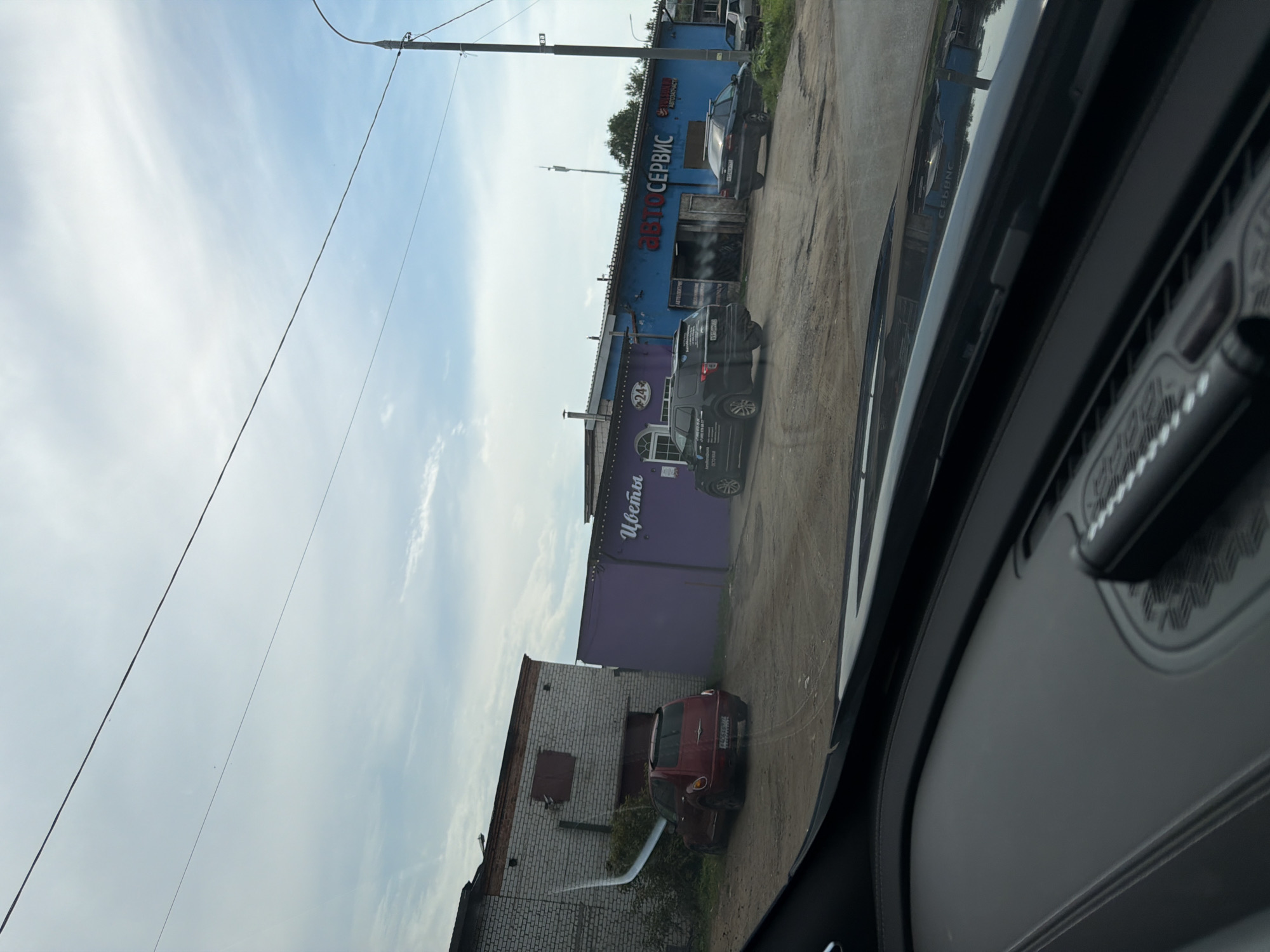 This is a street-level, slightly elevated, eye-level shot captured from inside a vehicle, looking out at a small commercial strip in Povarovo, Russia. The foreground is dominated by the dark interior of the car, specifically the dashboard and windshield. Beyond the windshield, a dusty, unpaved lot separates the car from a row of buildings.
To the left, a low-slung, pale brick building with a corrugated metal roof extension has a red PT Cruiser parked in front of it. The building to its right is painted a vibrant purple and features white lettering that appears to spell out "Цветы" (Flowers) and a "24" in a circle. Next to the purple building is a longer, blue building with red lettering that reads "АВТОСЕРВИС" (Auto Service), indicating a car repair shop. Several cars are parked in front of or near the auto service building, including a dark SUV and a dark sedan. The sky above is overcast with scattered clouds, and a utility pole with wires runs along the right side of the frame. The environment appears somewhat industrial and utilitarian.