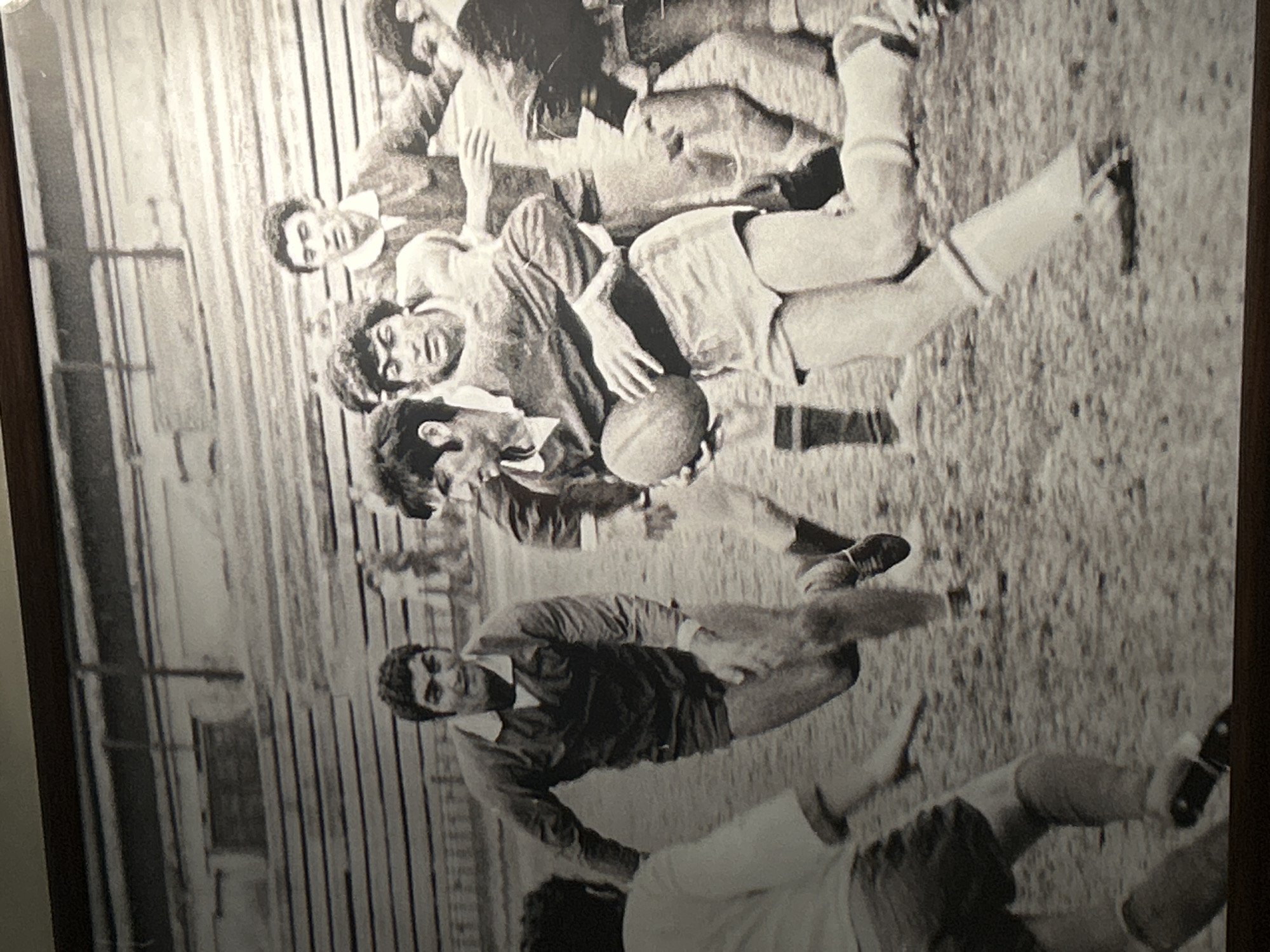 This black and white, action-oriented photograph captures a dynamic moment during a rugby or similar team sport game. The image features multiple male players, likely young adults, engaged in intense physical play on an outdoor field.
In the center of the frame, one player wearing a dark collared jersey and shorts is seen holding an oval-shaped ball, characteristic of a rugby ball. He is surrounded by other players, some in lighter-colored jerseys, indicating two opposing teams. Their body language and positions suggest a scramble for the ball or a tackle in progress, with players bending, reaching, and pushing against each other. Faces show concentration and effort.
The setting appears to be a sports pitch, possibly grassy or dirt, under natural daylight conditions. In the background, vertical structures, resembling fencing or spectator stands, indicate a designated sports venue. The monochrome format suggests the photograph is historical, likely from several decades ago, capturing an athletic event. There is no visible text in the image. Given the context, this scene likely depicts a rugby match in Yerevan, Armenia.