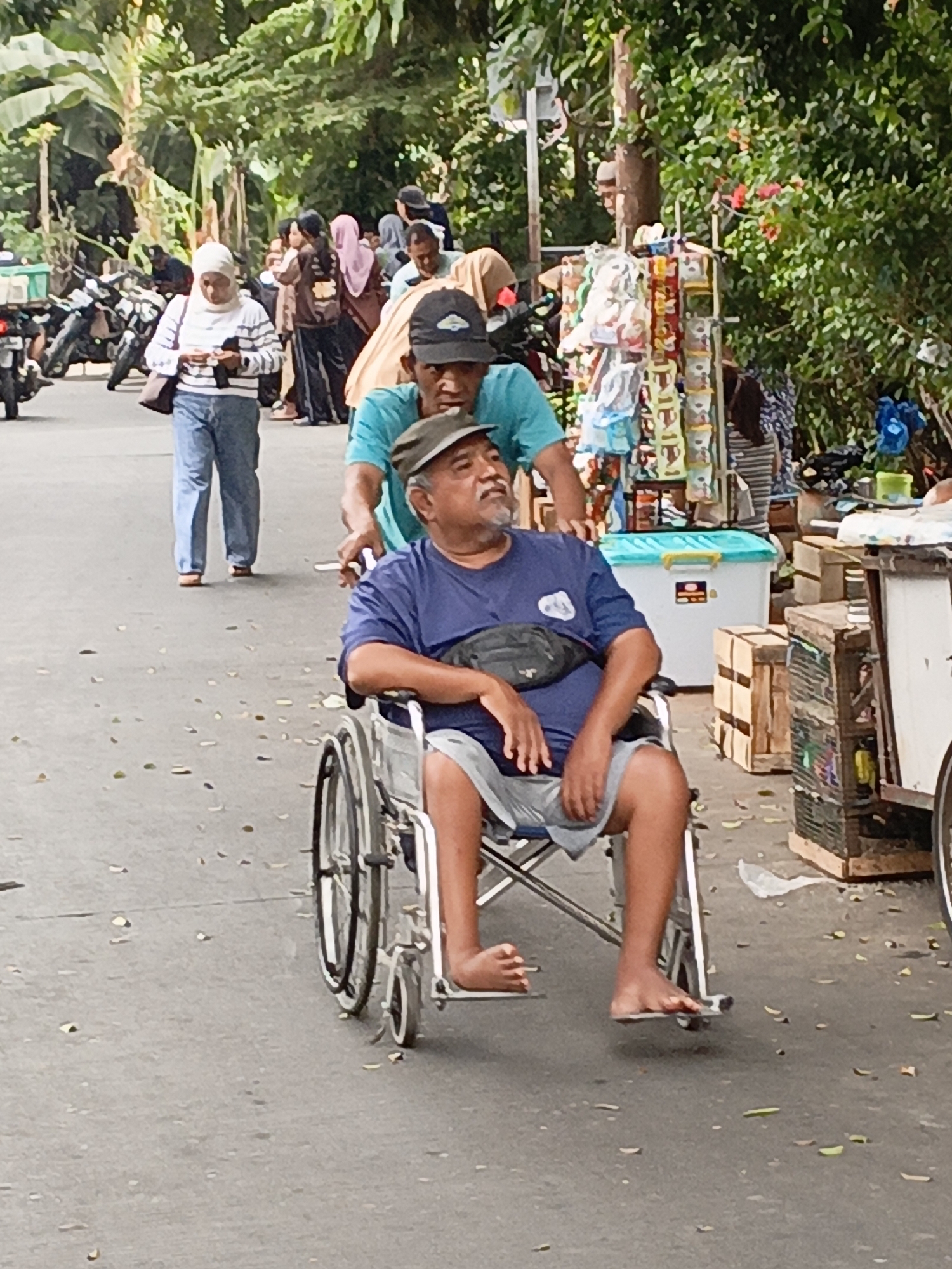 In East Jakarta, Indonesia, on an asphalt road with scattered leaves, a man sits in a wheelchair being pushed by another man from behind. The man in the wheelchair wears a blue t-shirt and grey shorts, with a black fanny pack slung across his chest. The man pushing the wheelchair wears a teal t-shirt and a dark baseball cap. In the background, a woman in a white hijab and striped long-sleeved shirt walks away from the camera, looking at her phone. Other pedestrians and motorbikes are visible further in the background, along with stalls selling various goods. The environment is characterized by lush green trees and foliage.