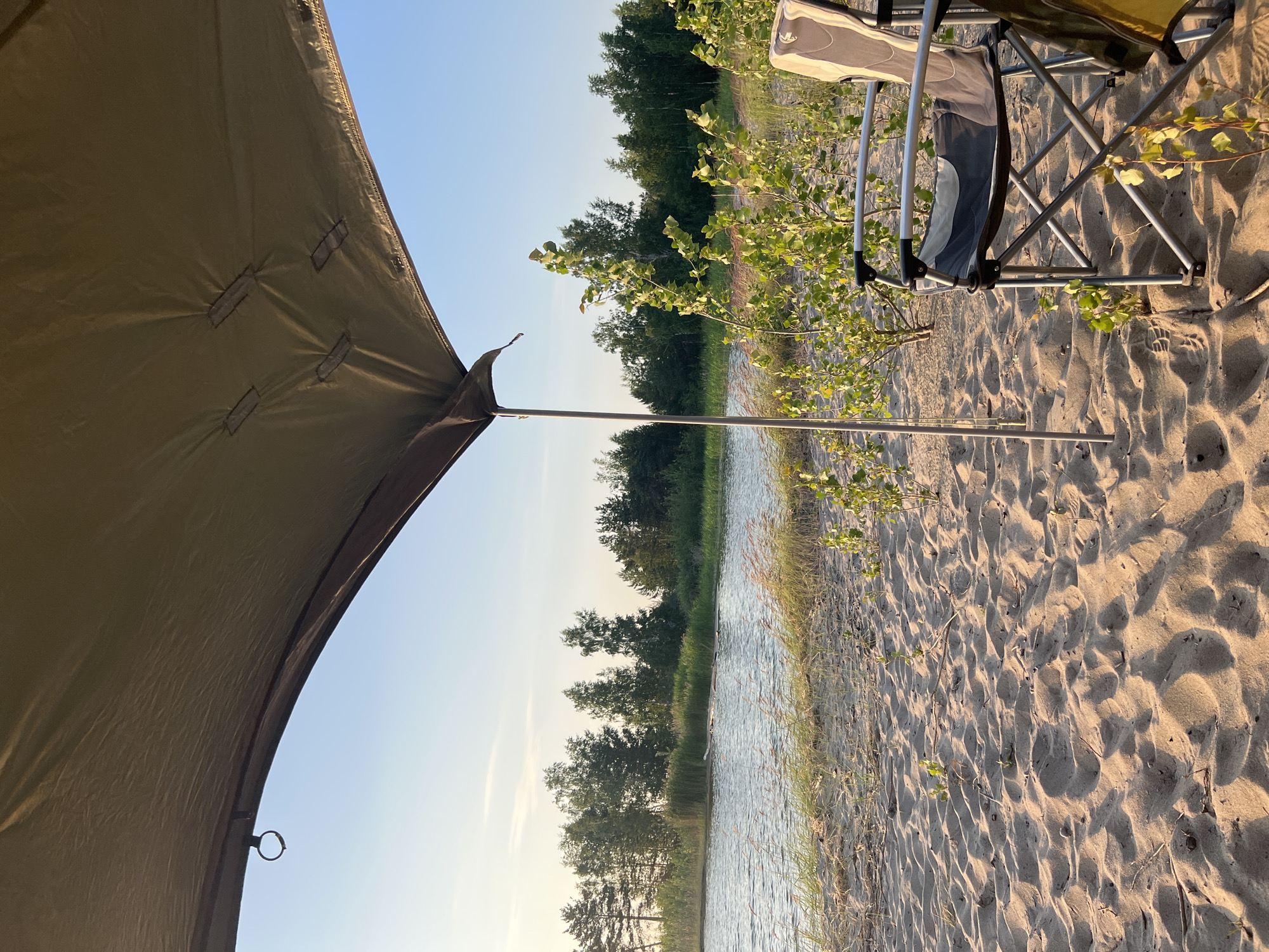 This is a ground-level, outdoor photograph taken on a sandy beach next to a body of water. The perspective is from under a canvas awning, which extends from the top left to the top right of the frame. The awning is a muted olive green color and appears to be part of a camping setup, with a metal pole supporting it in the center of the image.
In the foreground, the sandy beach is textured with footprints and small divots. Scattered across the sand are a few dry twigs and small, leafy green plants. To the right, a silver and black folding camp chair is visible, angled towards the water. Part of another chair or bag is visible to the right of the primary chair.
The middle ground features a gently rippling body of water, reflecting the clear, pale blue sky. On the far side of the water, a dense line of trees forms the horizon. The trees are a mix of evergreens and deciduous species, with some taller pines and lighter-colored birches. The shoreline on the other side of the water is lined with taller reeds or marsh grass. The lighting suggests it is late afternoon or early evening, with soft sunlight casting shadows.
There are no people or animals visible in the frame. The location is identified as Borovoye, Russia, which is known for its numerous lakes and forested areas, consistent with the scenery depicted.